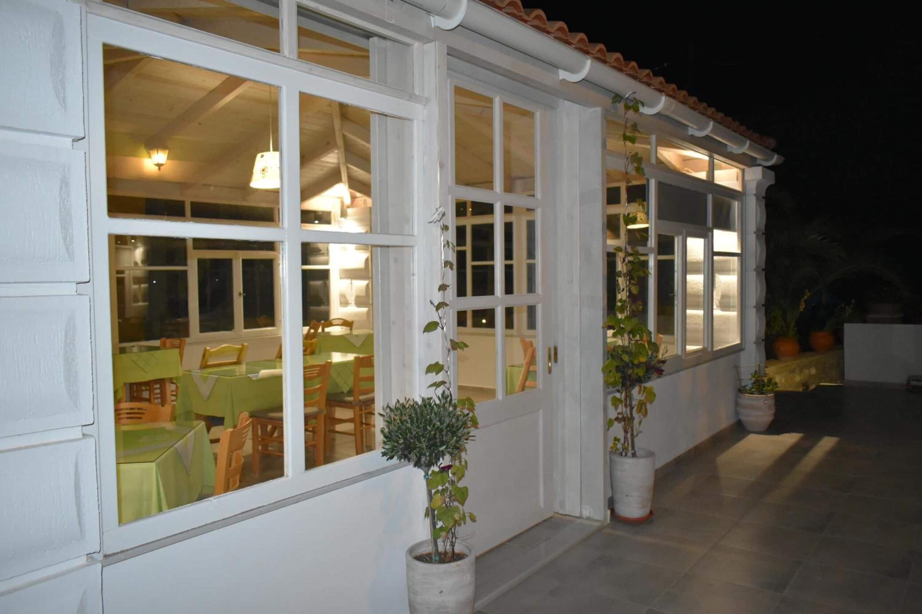 A covered outdoor dining area with multiple tables and chairs, large windows, and potted plants.