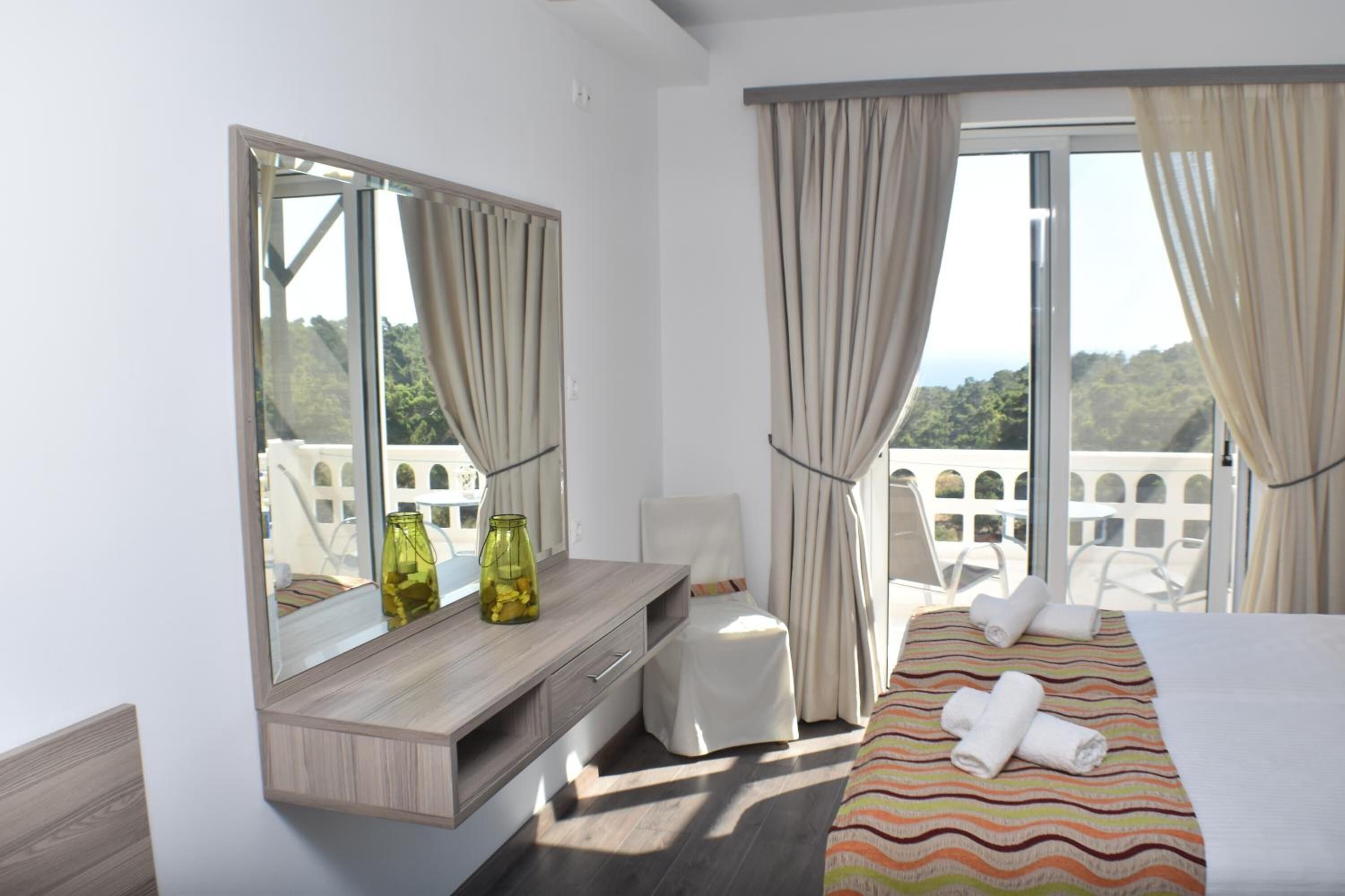 Habitación de hotel con cama, tocador con espejo, una pequeña mesa con un florero y un balcón con sillas.