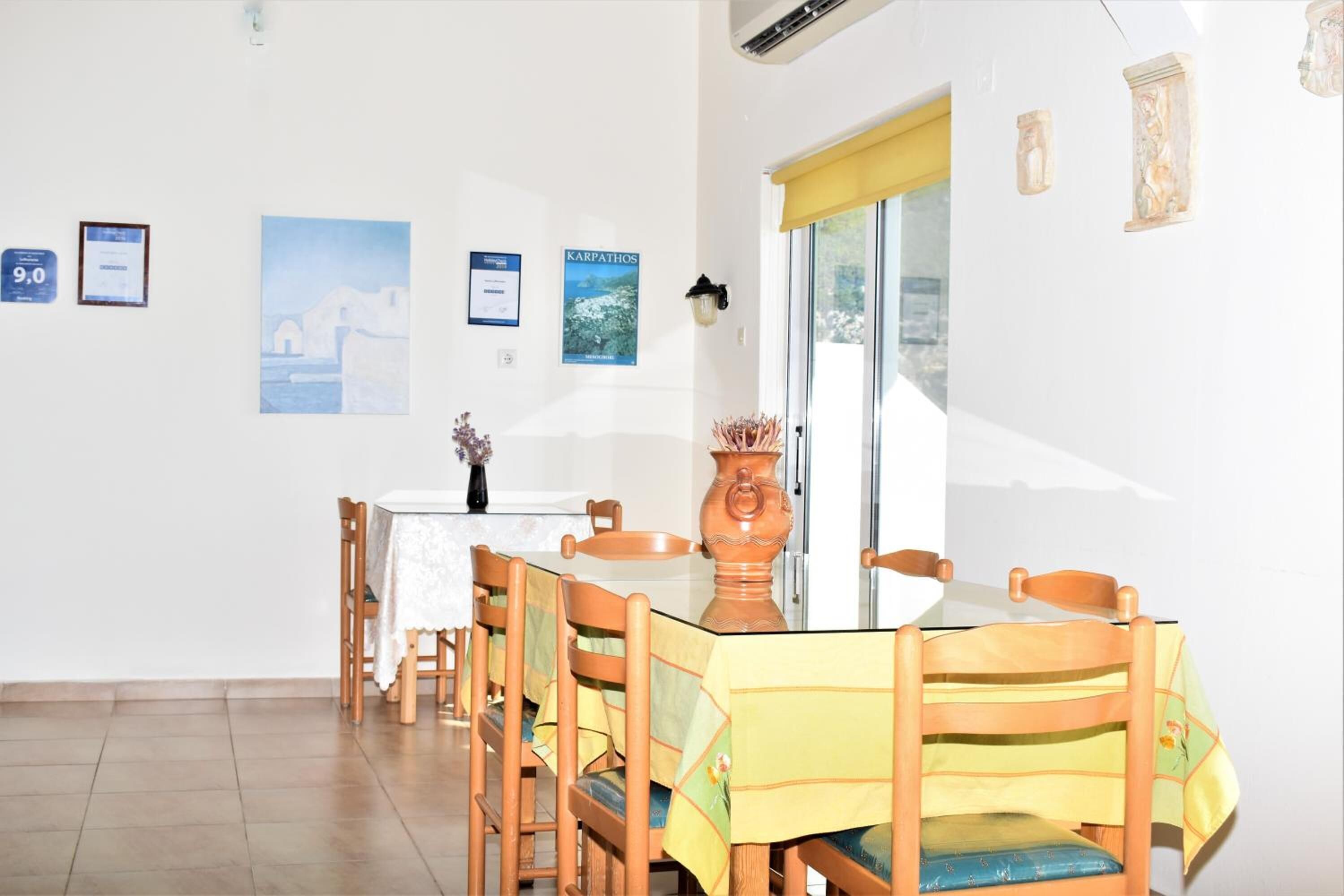 Un coin repas avec une table dressée pour quatre personnes, entouré de chaises en bois recouvertes de nappes jaunes. Des certificats encadrés ornent le mur, et une grande fenêtre est dotée d’un store jaune.