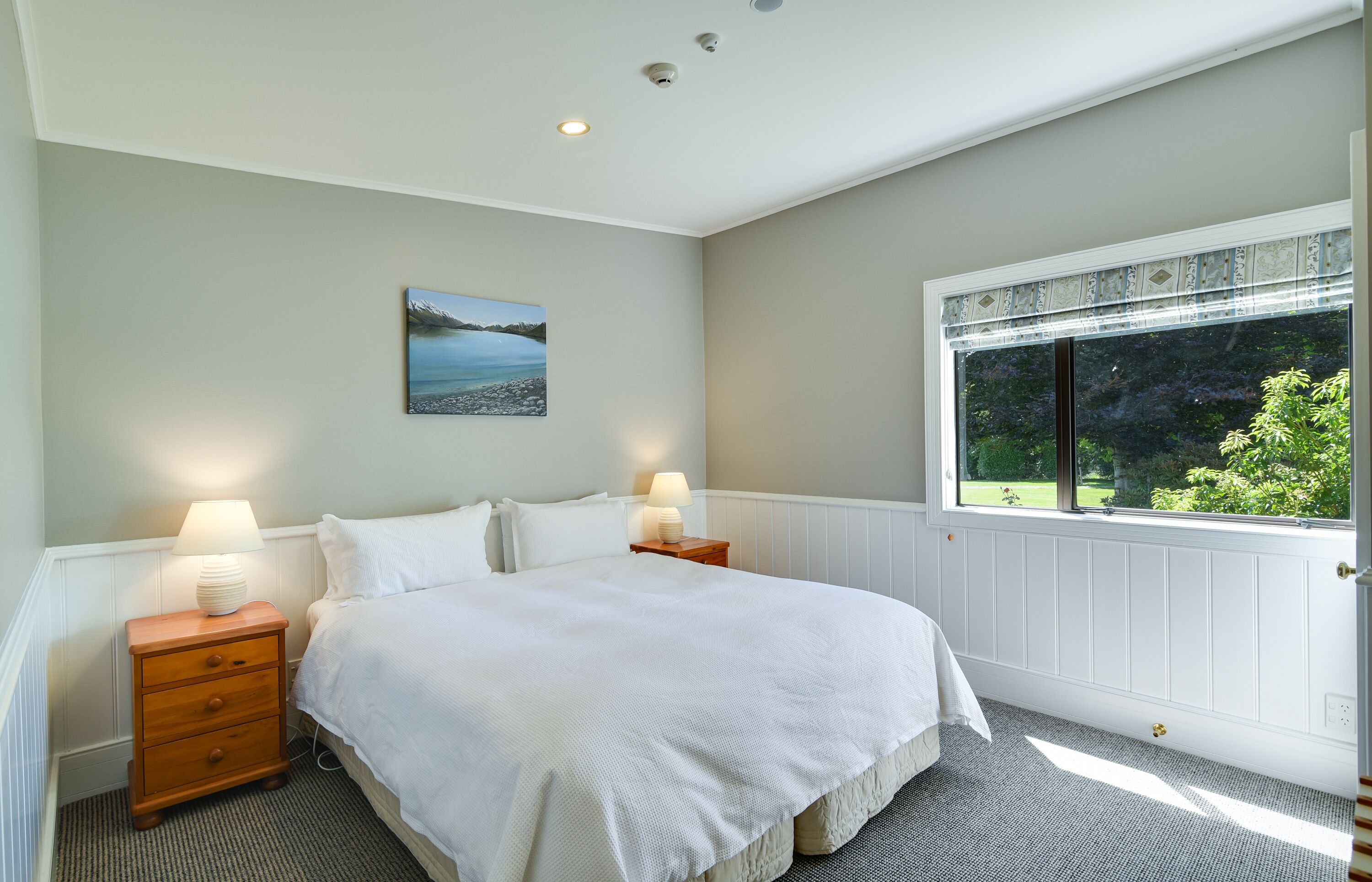 A bedroom with a large bed, two bedside tables with lamps, a window with a view of greenery, and a painting on the wall.