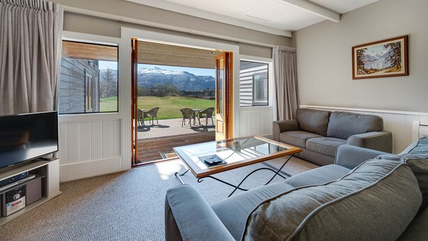 A living room with a large TV, a glass coffee table, a grey sofa, and a view of a mountain landscape through open doors. - Manata Lodge (Lower Shotover)