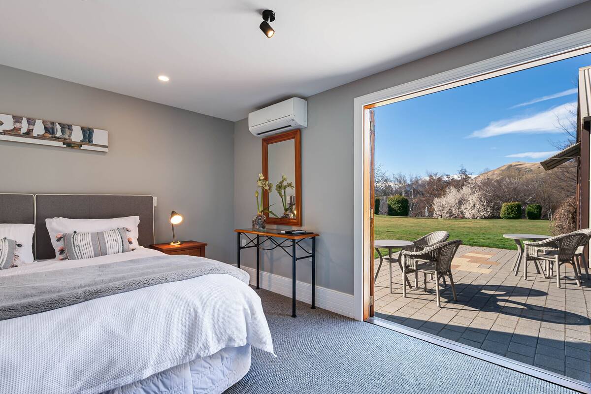 A bedroom with a large bed, bedside table, lamp, and a view of a patio with outdoor furniture.