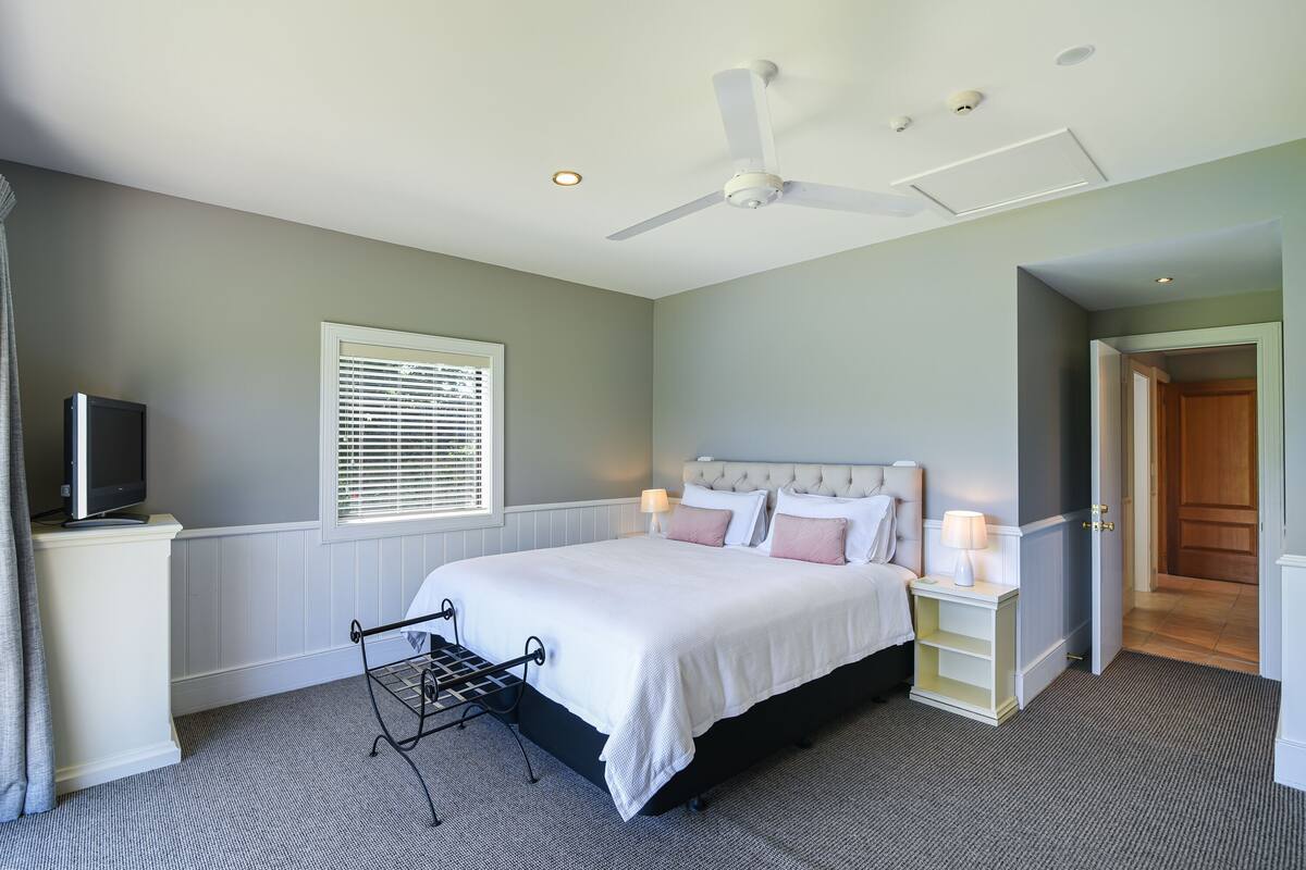 A bedroom with a bed, bedside tables, a ceiling fan, a TV, and a window with blinds.