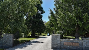A gravel driveway leading to a gated entrance with a stone wall and a sign reading "Manata Lodge." - Manata Lodge (Lower Shotover)