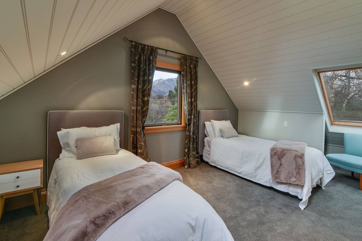 A bedroom with two beds, a window with curtains, a nightstand, and a view of mountains.