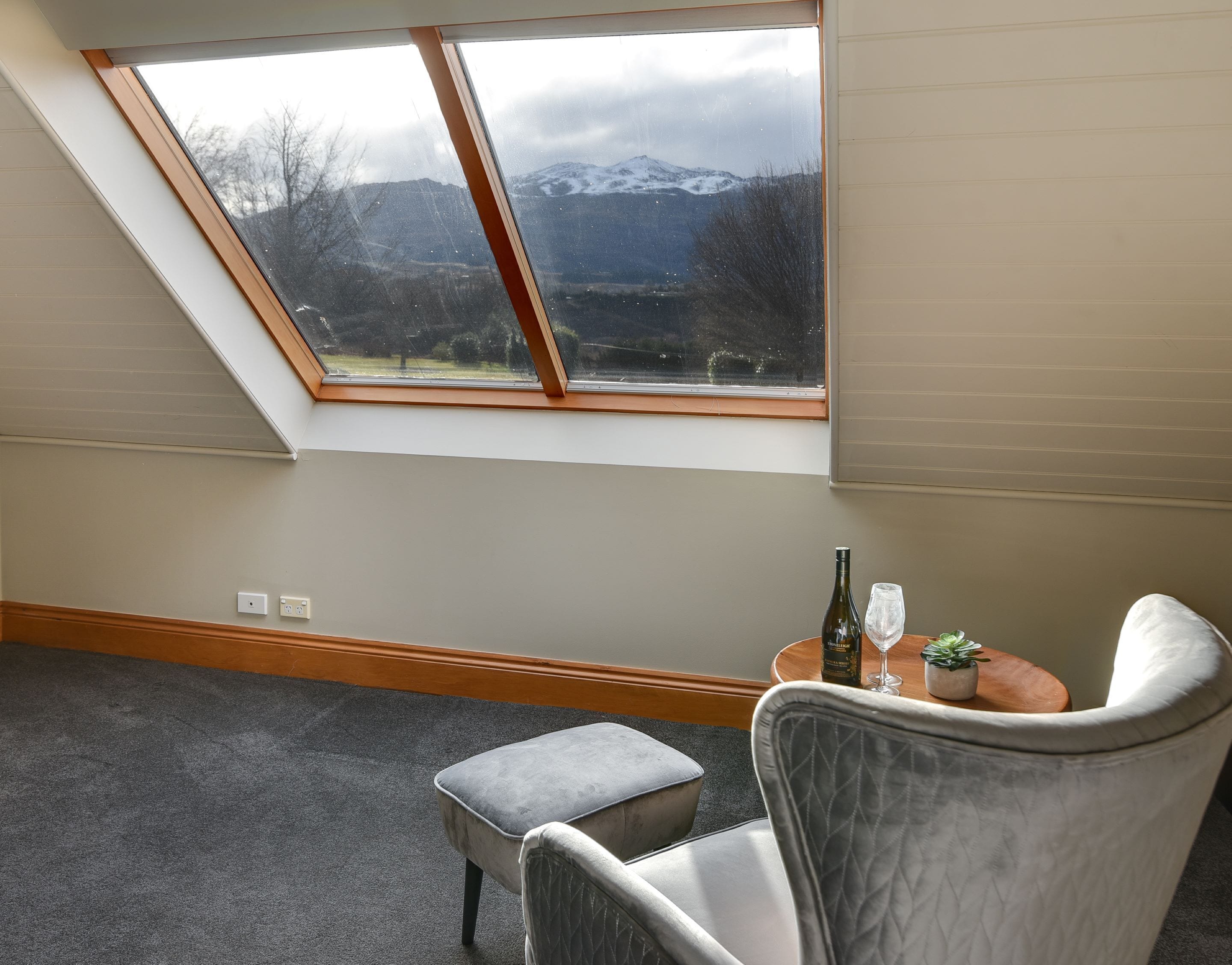 A room with a sloped ceiling, a skylight, a grey cushioned chair, a small table with a bottle and glass, and a view of a snowy mountain.