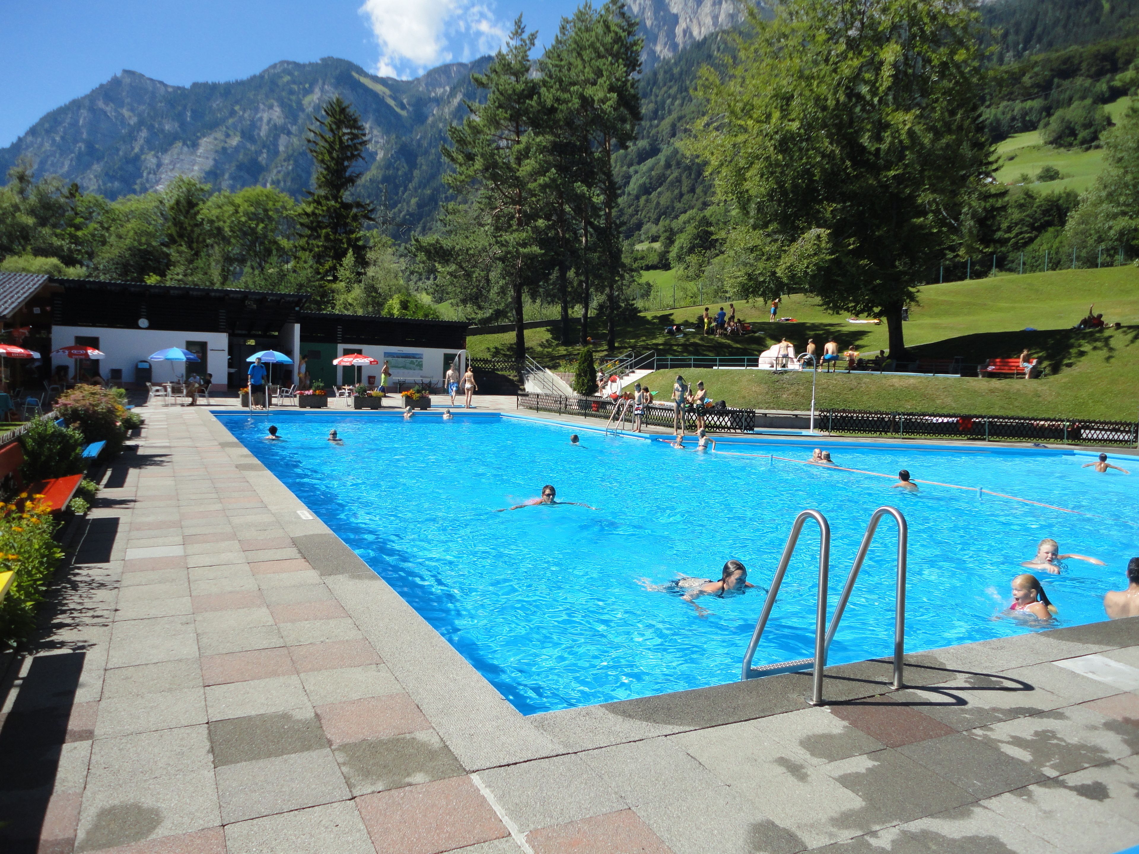 Una piscina con gente nadando y tomando el sol, rodeada de montañas y vegetación.