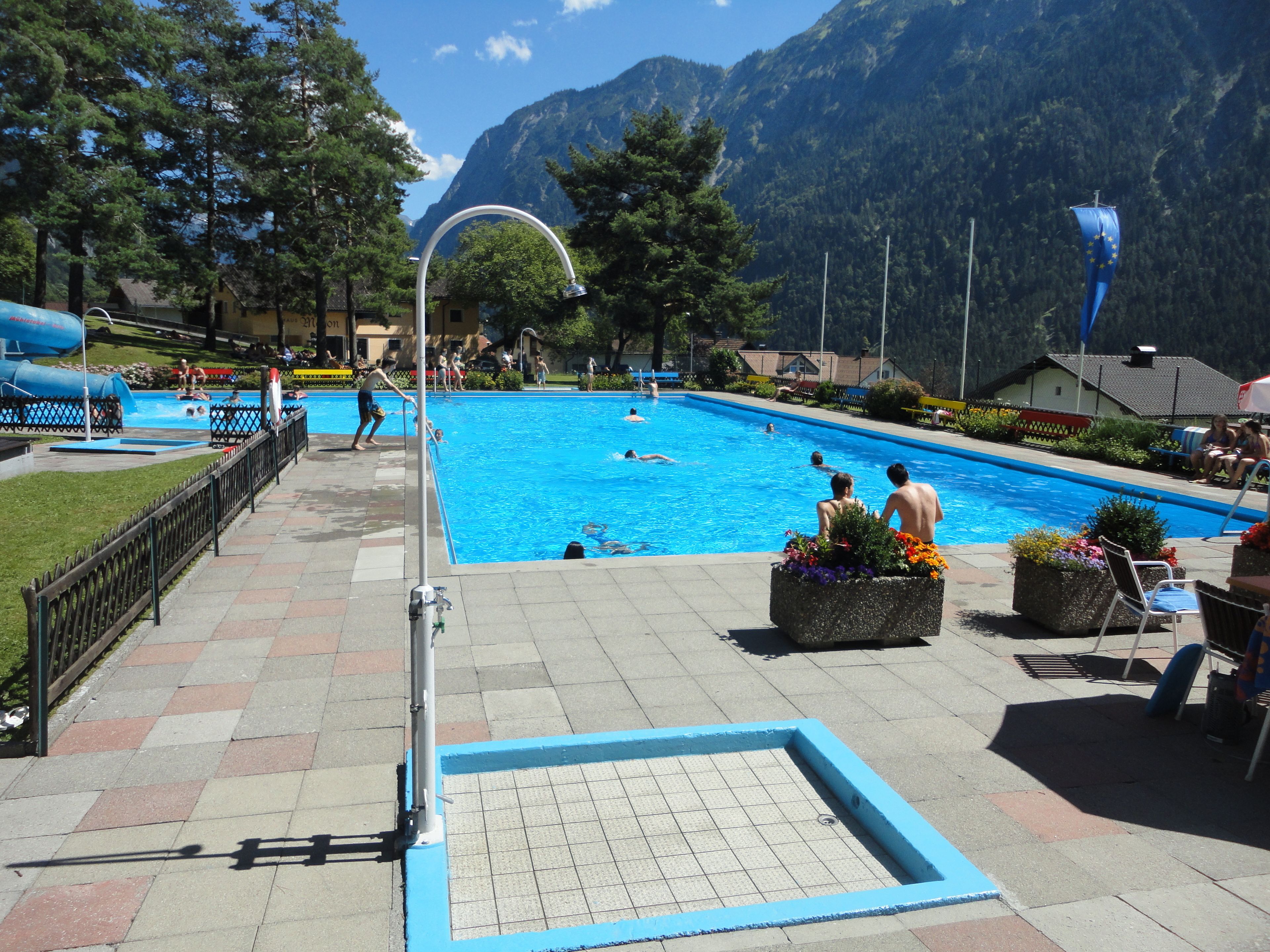 A swimming pool with people swimming and playing, surrounded by mountains and trees.