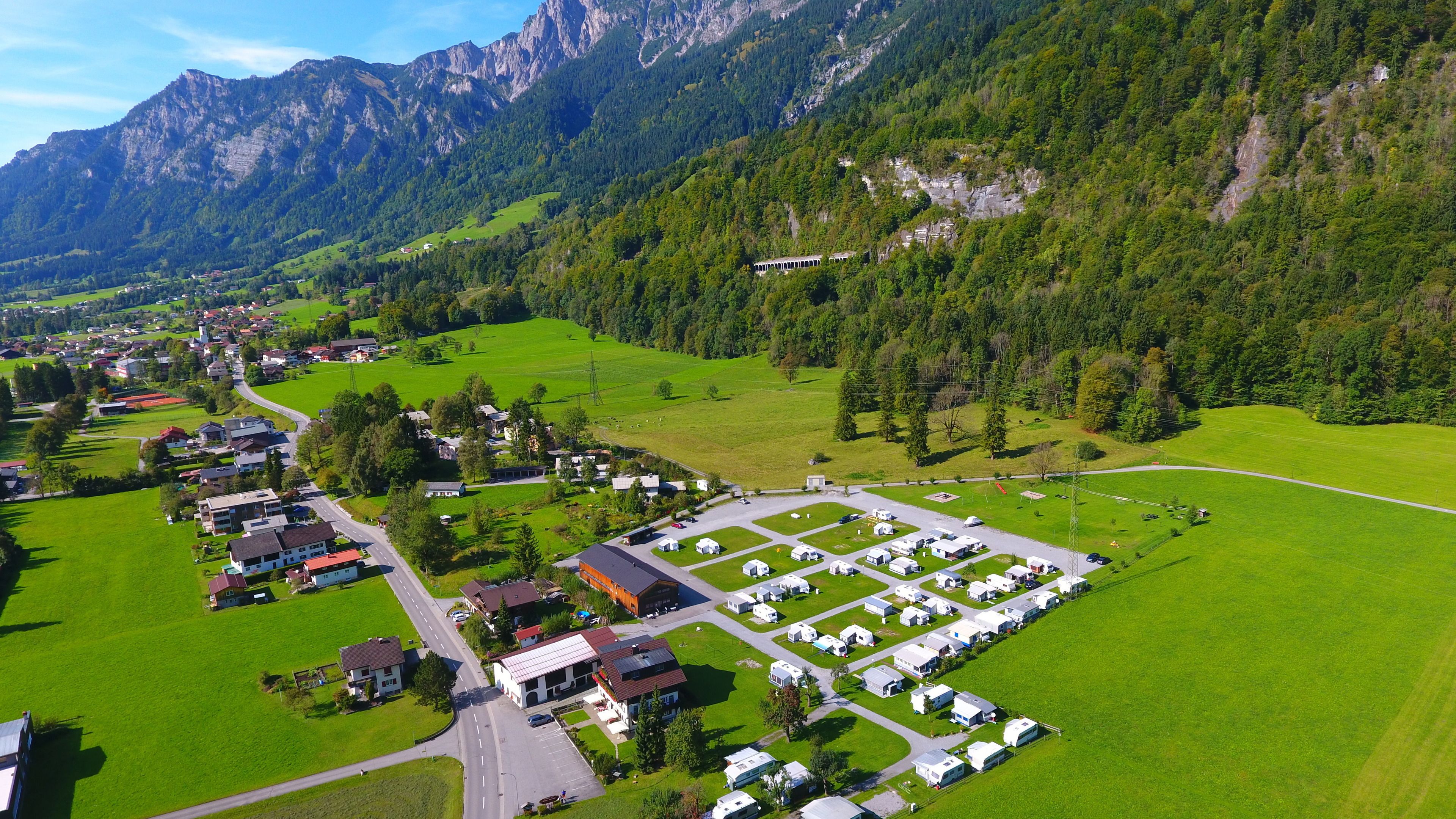 Un terrain de camping avec des emplacements pour camping-cars, un parking et des bâtiments situés dans une vallée, avec des montagnes en arrière-plan.