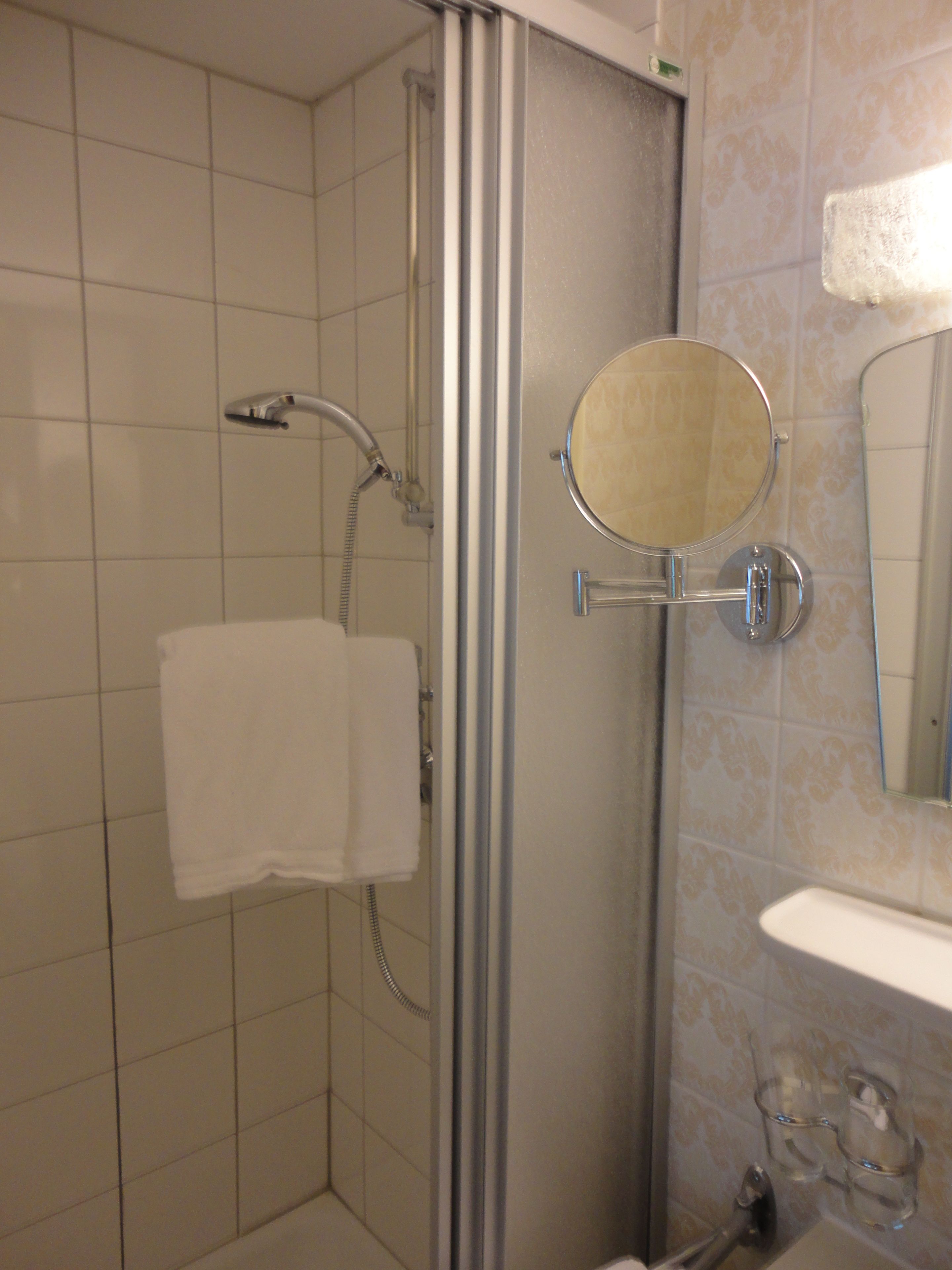 A bathroom with a glass shower enclosure, a white towel hanging on the wall, a round mirror, and a sink with two glasses.