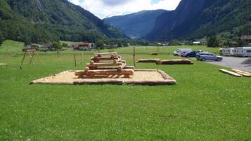 A grassy field with a wooden structure in the center, surrounded by mountains and a few houses in the background.
