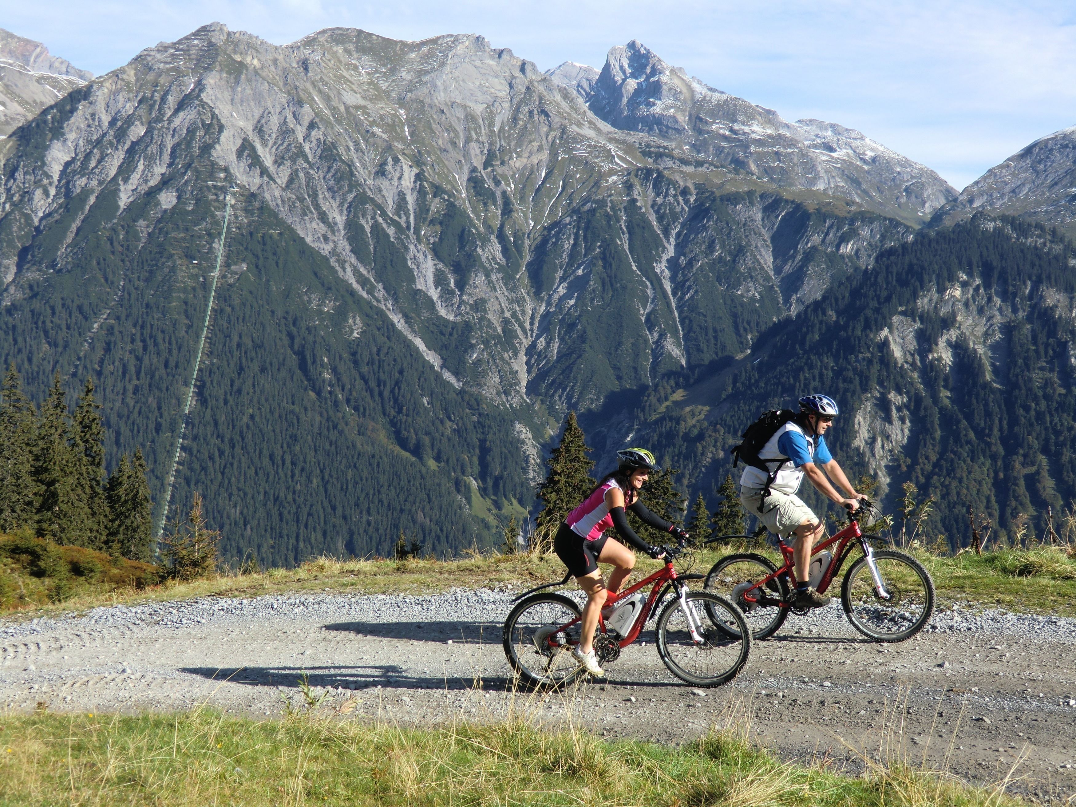 Two cyclists on a gravel path with a mountainous backdrop.