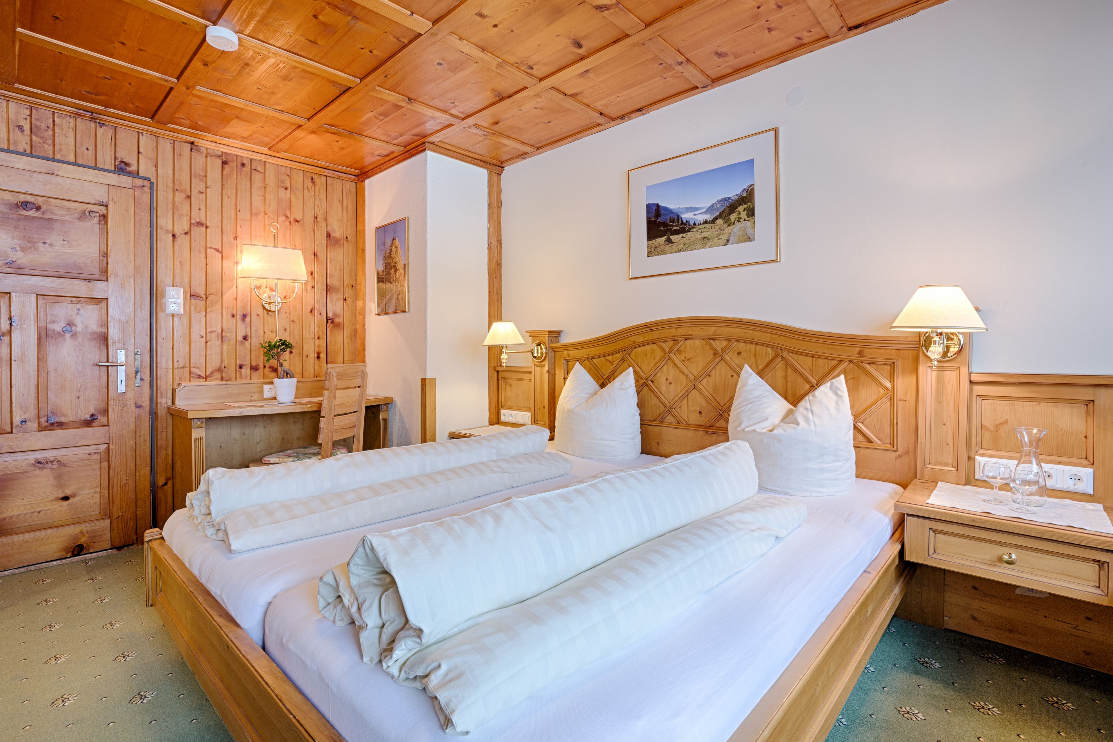 Cama doble con sábanas blancas, cabecera de madera, dos lámparas de noche y una mesita de noche con un florero.
