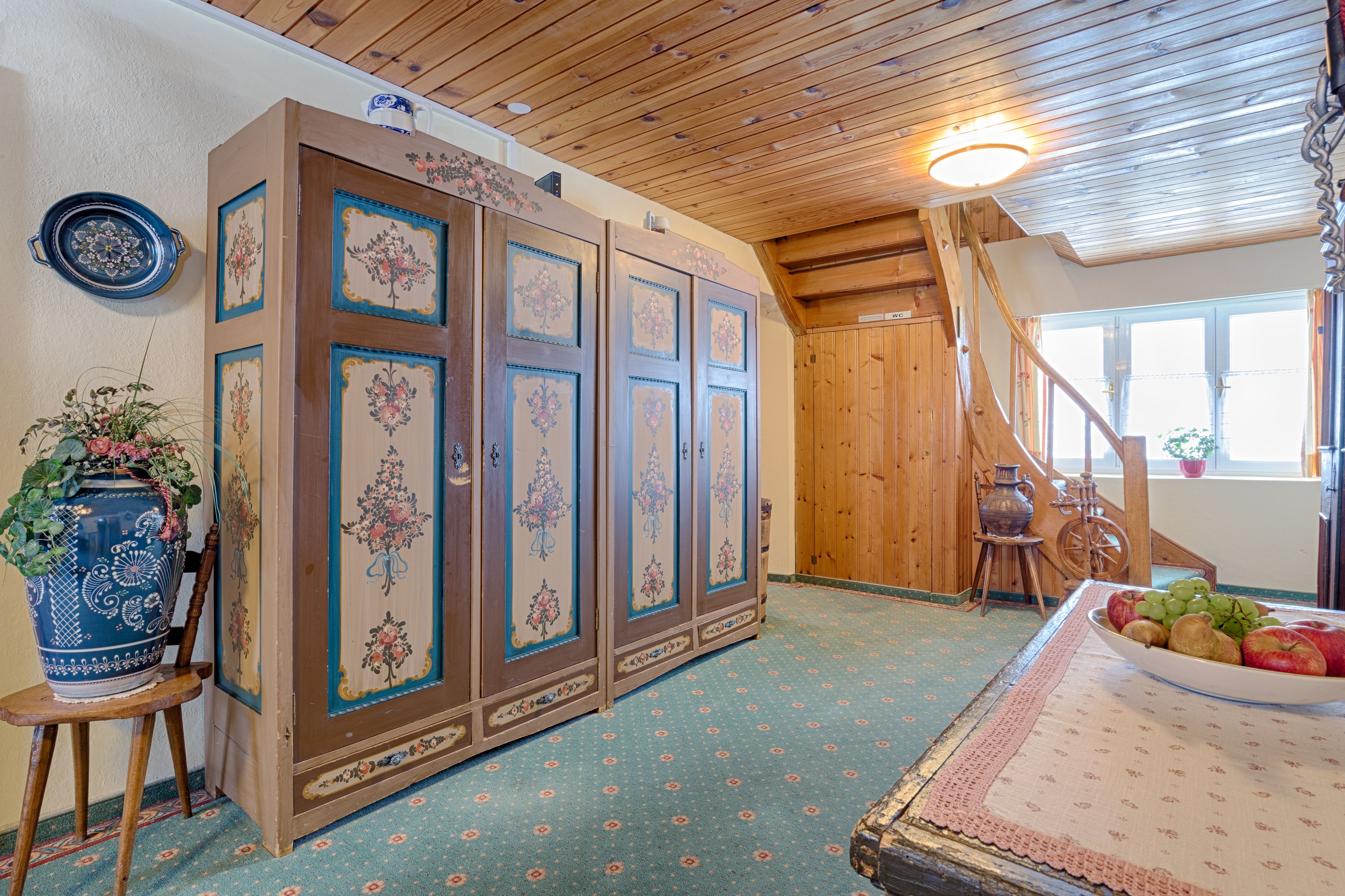 A room with a wooden ceiling, a large cabinet with floral designs, a vase with flowers, and a table with fruits.