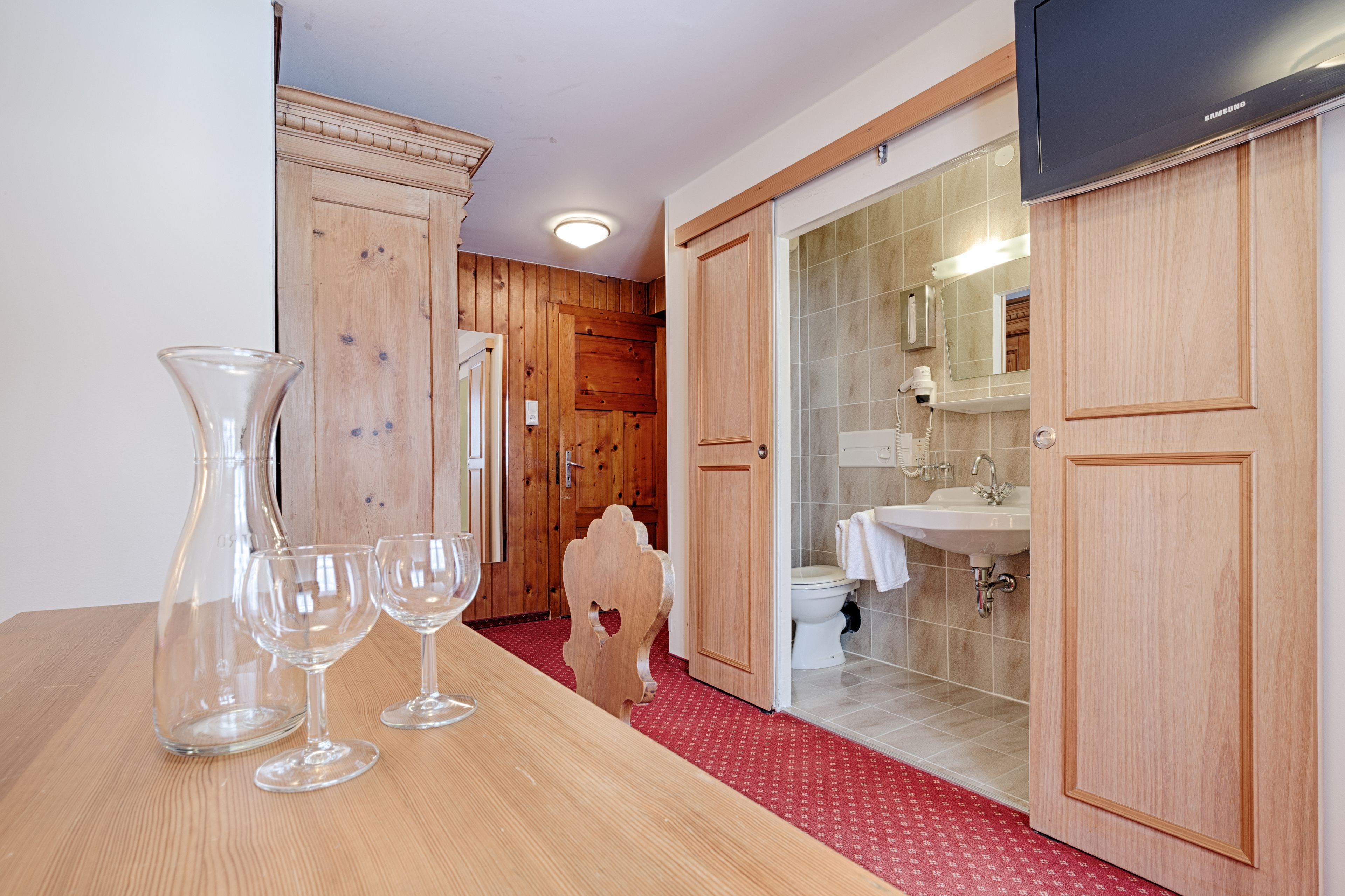 Una mesa de madera con una botella de vidrio transparente y dos copas de vino, un mueble de madera y un baño con un lavamanos y un inodoro visibles a través de una puerta abierta.