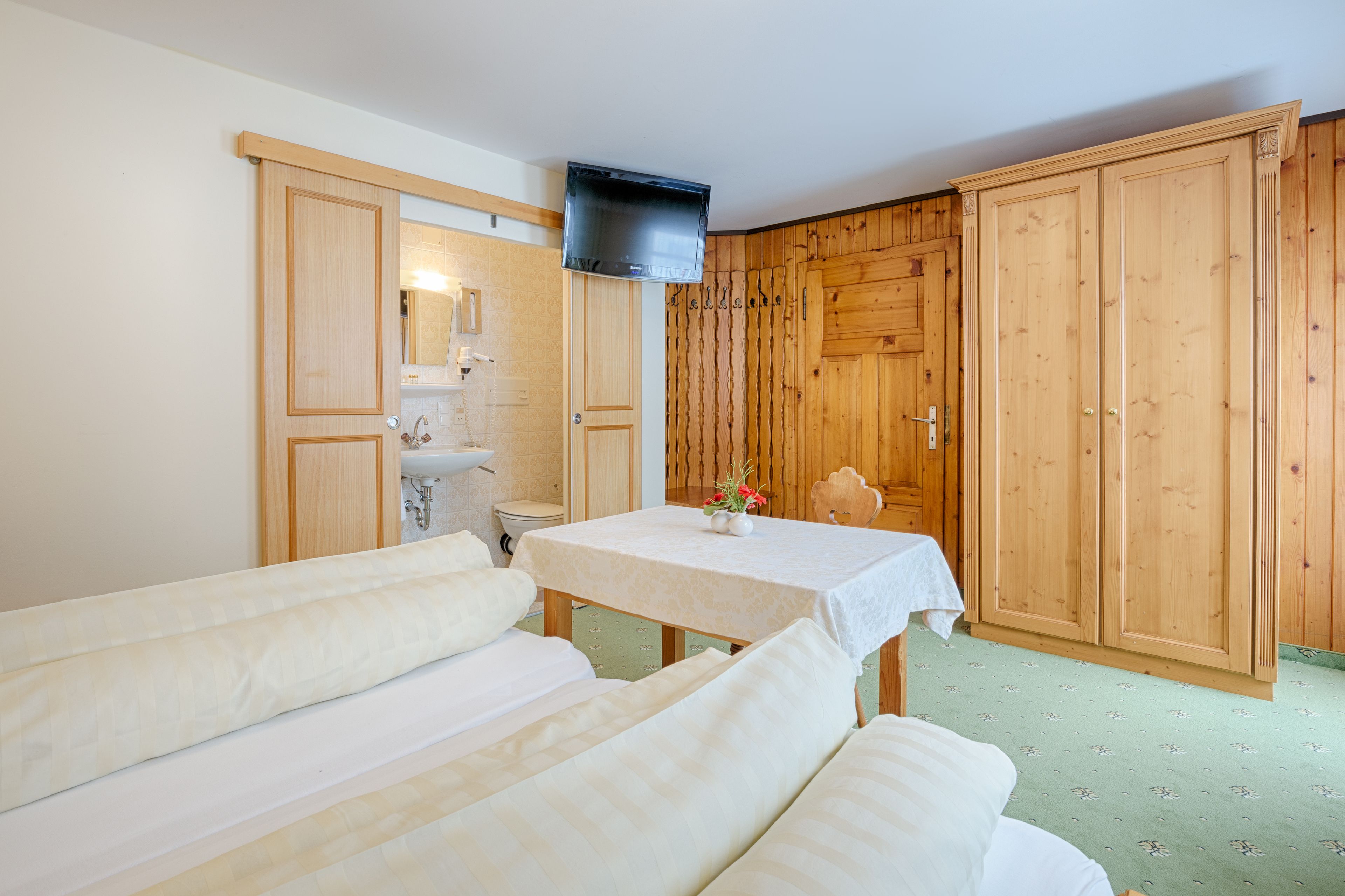 Quarto com armários de madeira, televisão fixada na parede, mesa com toalha branca e duas camas com roupa de cama branca.
