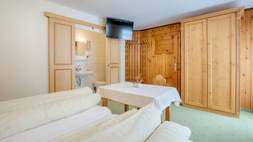 Quarto com armários de madeira, televisão fixada na parede, mesa com toalha branca e duas camas com roupa de cama branca.