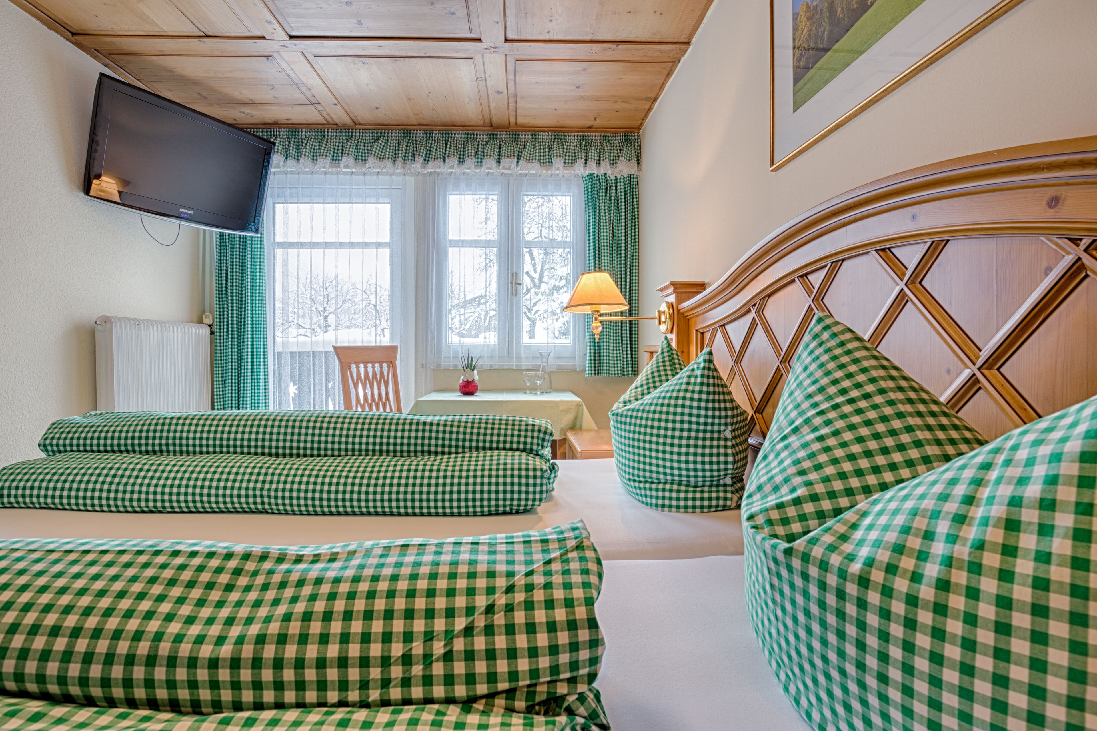 A cozy room with a checkered sofa set, a wooden headboard, a television mounted on the wall, and a window with curtains.