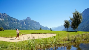 A golf course with a player in a white shirt and brown pants, surrounded by green grass, sand bunkers, and a mountain backdrop. - Landhaus Walch (Innerbraz)