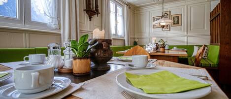 Un comedor con una mesa de madera preparada para dos personas, un banco con cojín verde, un reloj y una araña.