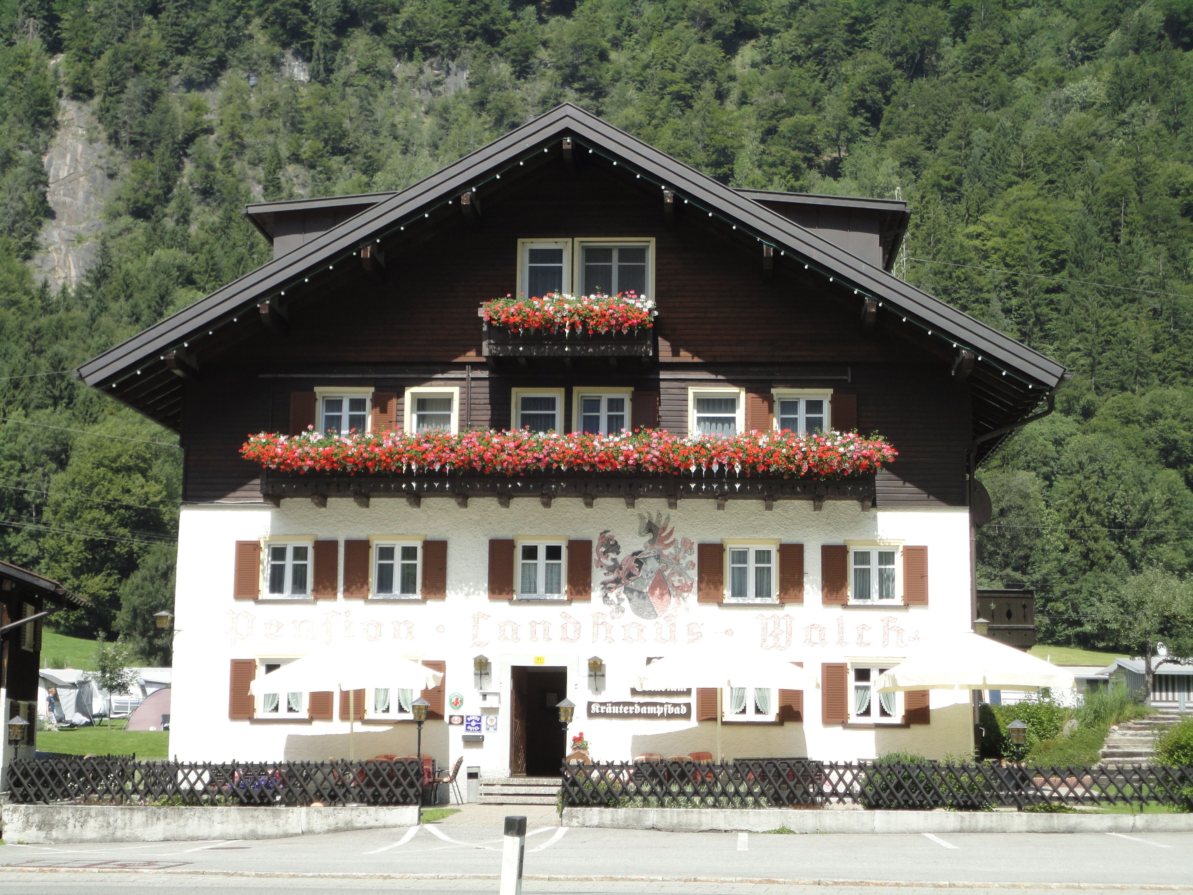 A traditional chalet with a balcony adorned with flowers, a sign indicating it is a restaurant, and a view of a forested mountain in the background.