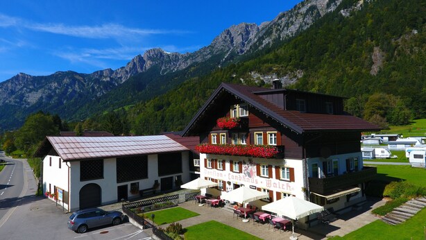 A traditional chalet-style building with a balcony adorned with red flowers, a paved outdoor seating area, and a view of a mountain range. - Landhaus Walch (Innerbraz)