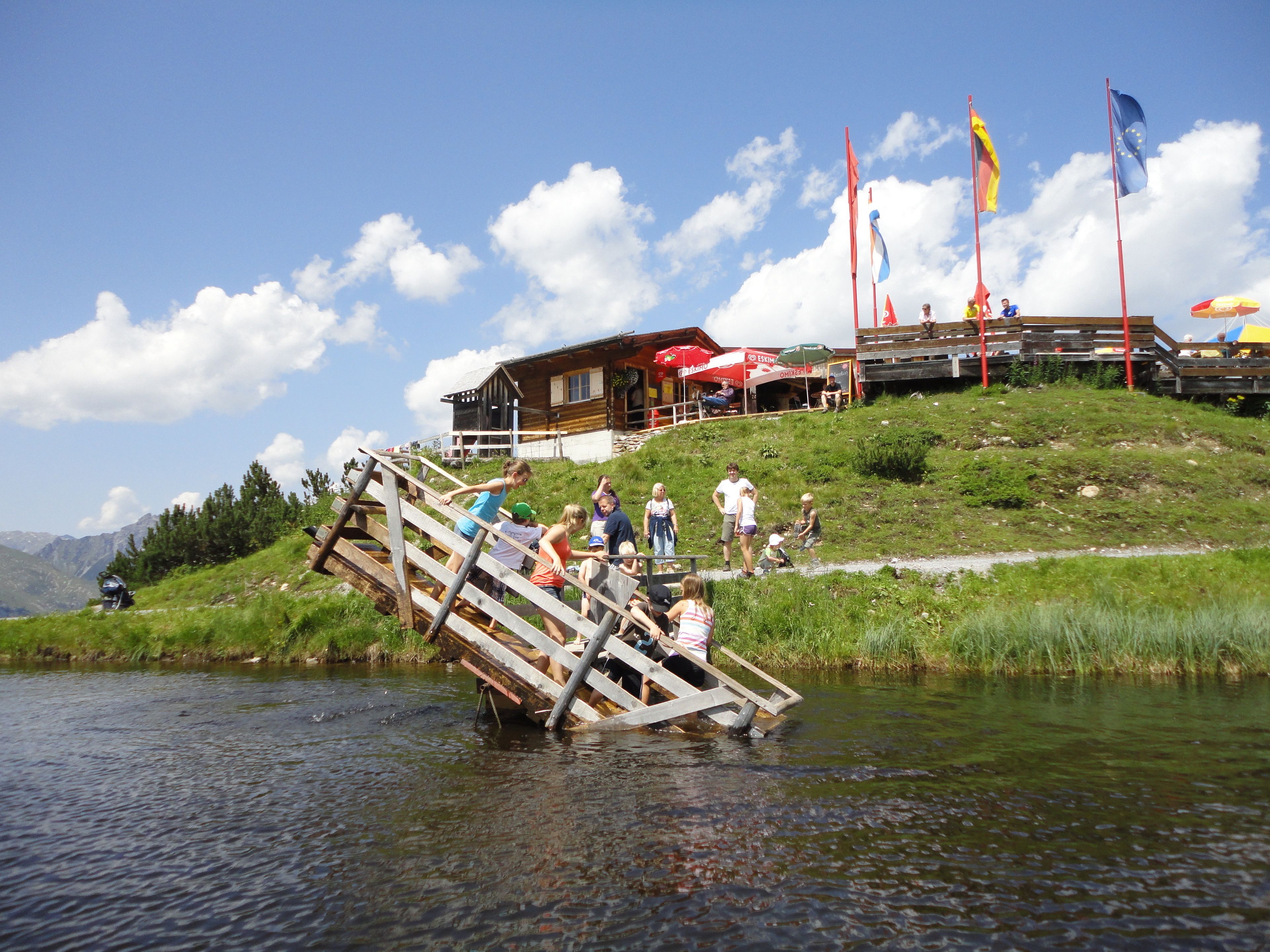A wooden bridge over a river with people crossing. A building with flags and outdoor seating is visible in the background.