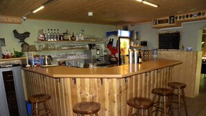 Una zona de bar con taburetes de madera, estantes con botellas y vasos, y un refrigerador.