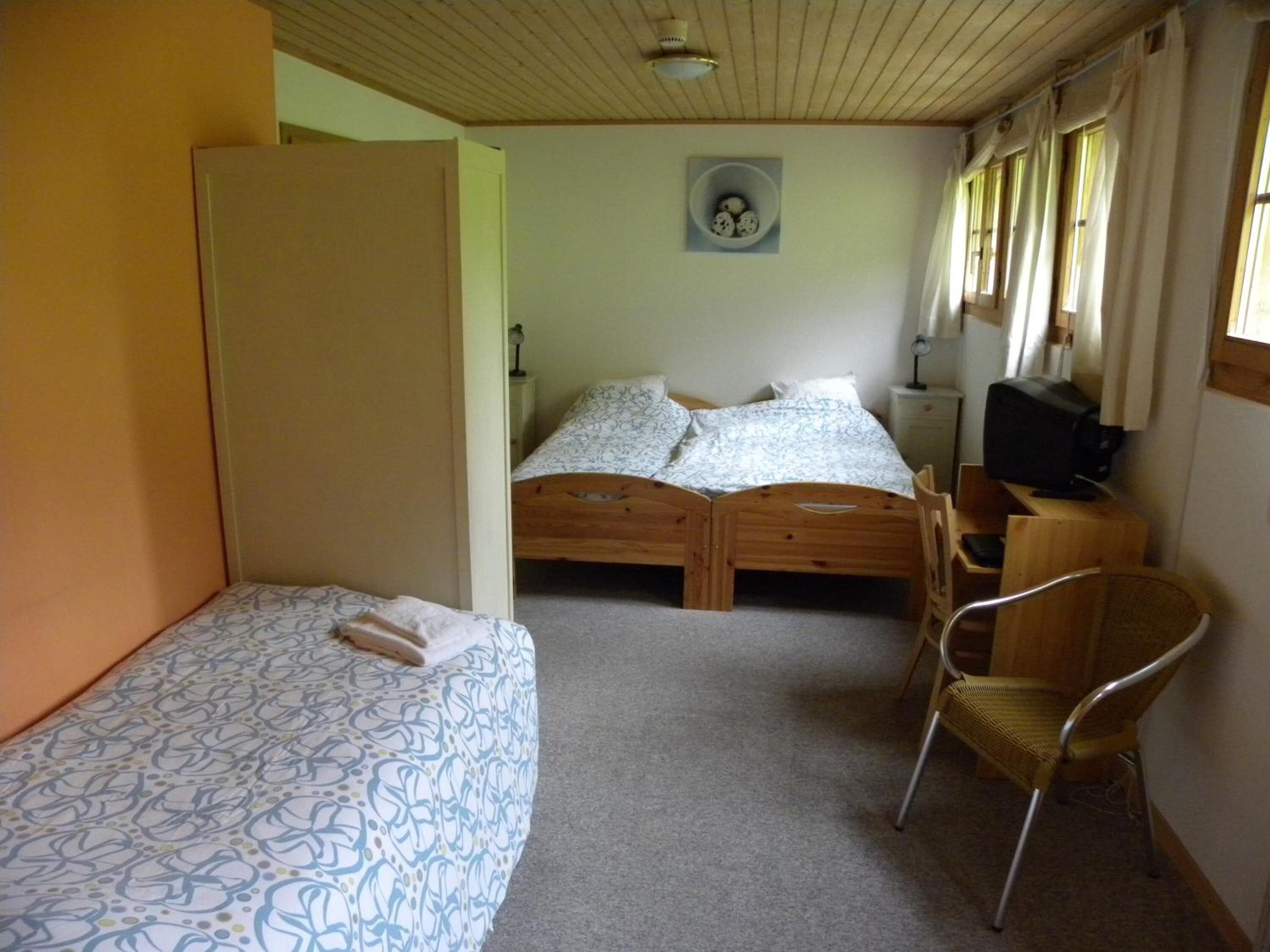 A room with two wooden beds, a chair, a desk with a computer, and a small wall-mounted shelf.