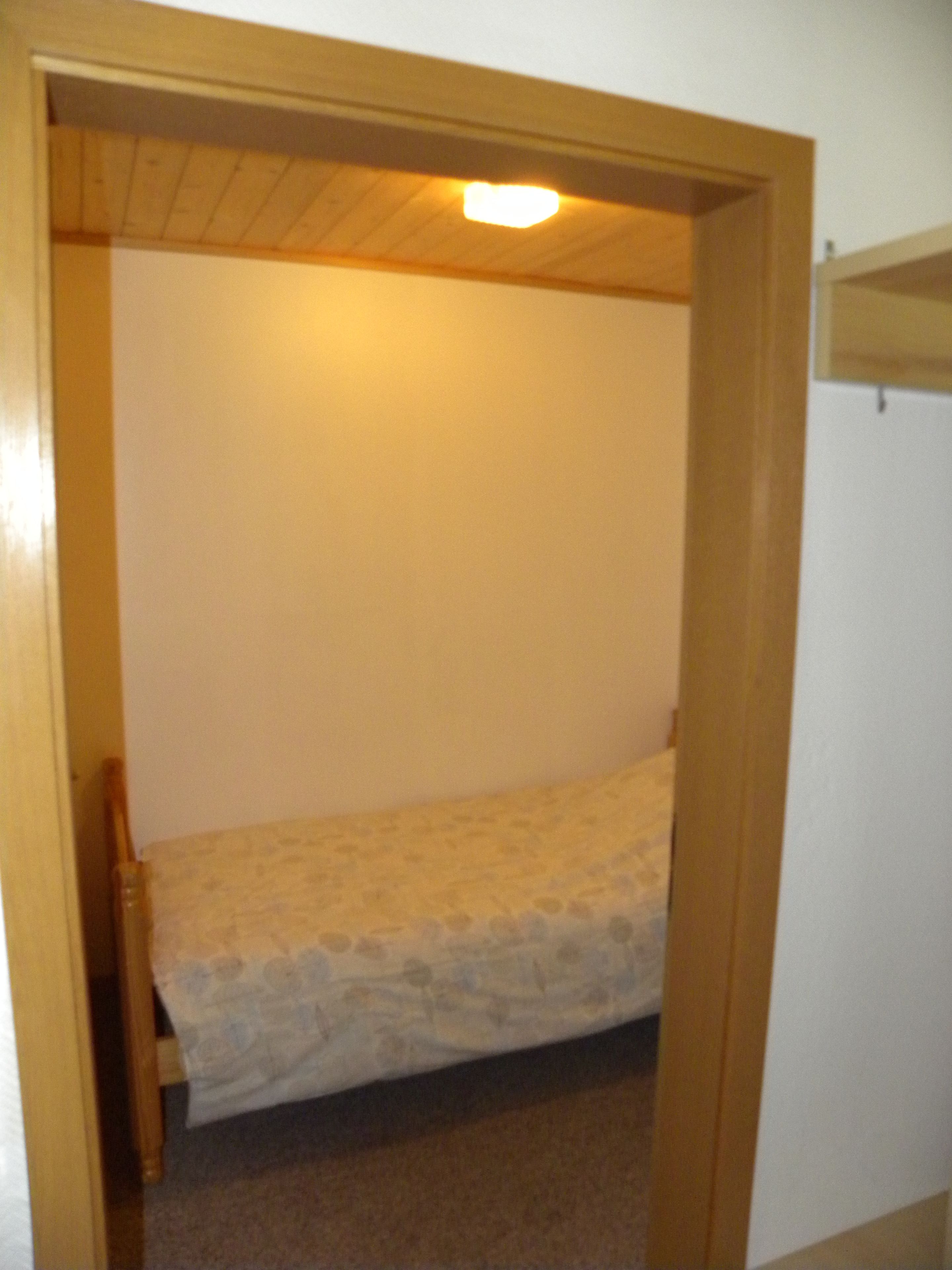 A small, narrow room with a single bed, a wooden door frame, and a light fixture on the ceiling.