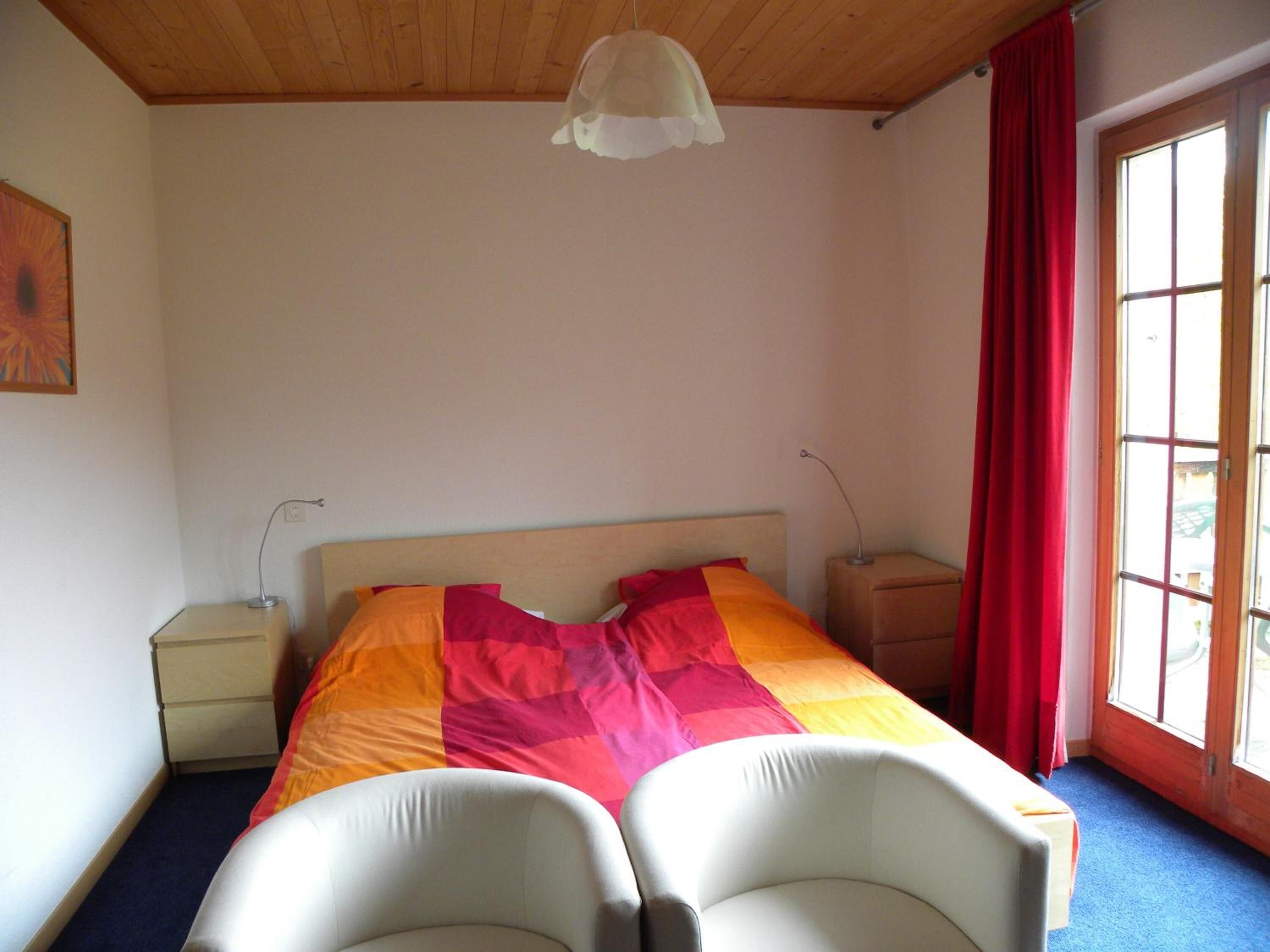 Ein Schlafzimmer mit einem Bett, zwei Nachttischen, einer Lampe, einem Fenster mit roten Vorhängen und einer Deckenleuchte.