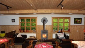 Una habitación acogedora con paredes y techo de madera, una estufa de leña central, una mesa de comedor con sillas y ventanas amplias con vegetación afuera.