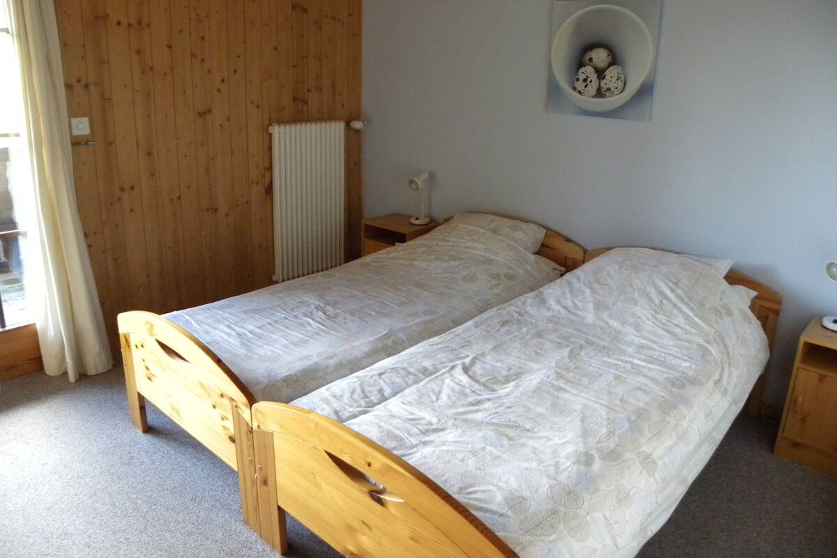A small, single-bed room with wooden walls and a radiator. There is a small bedside table with a lamp and a wall-mounted shelf with decorative items.