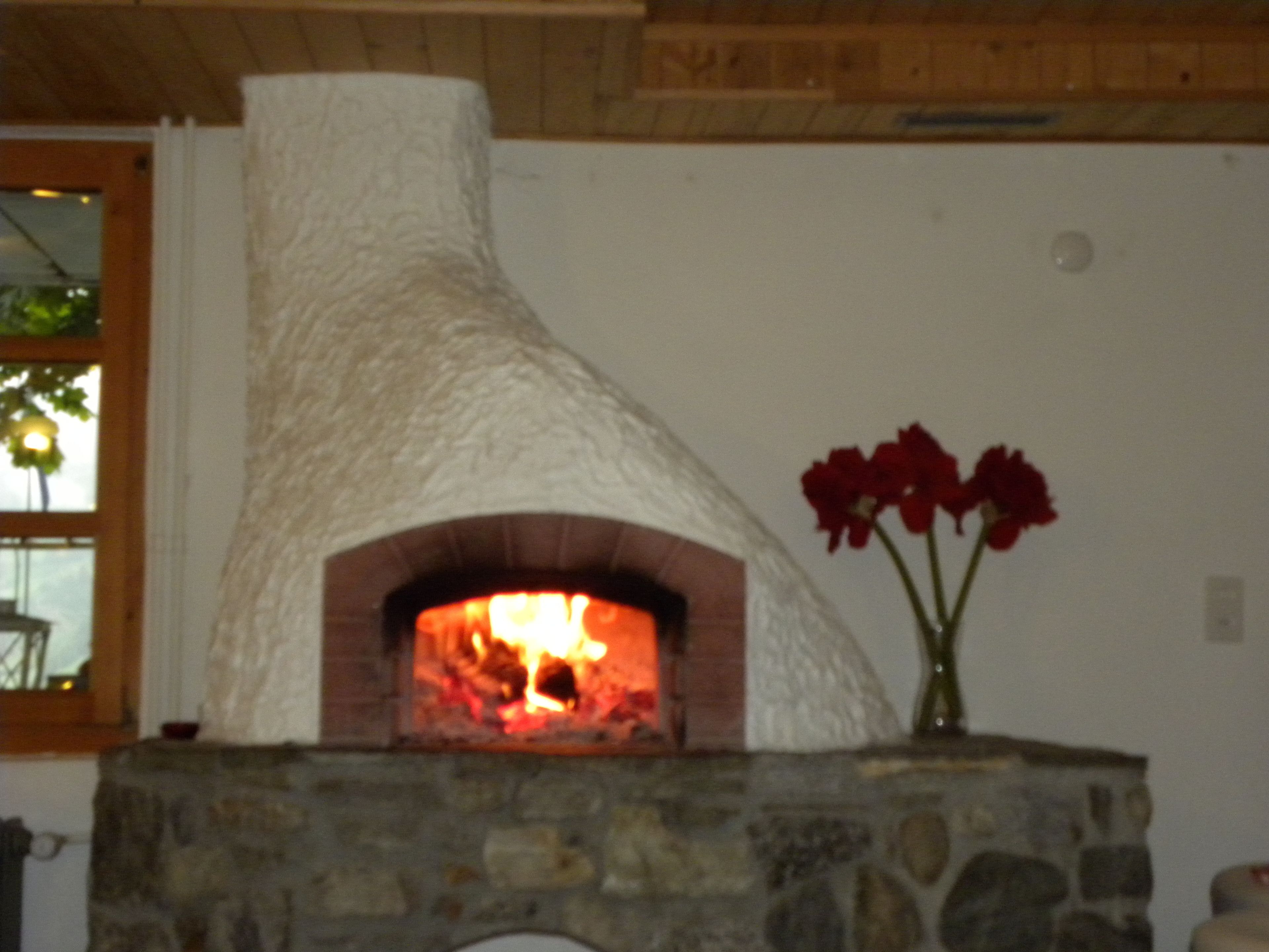 Une cheminée en pierre avec un feu qui crépite à l’intérieur, une cheminée blanche à la texture particulière et un vase rempli de fleurs rouges posé sur le manteau de la cheminée.