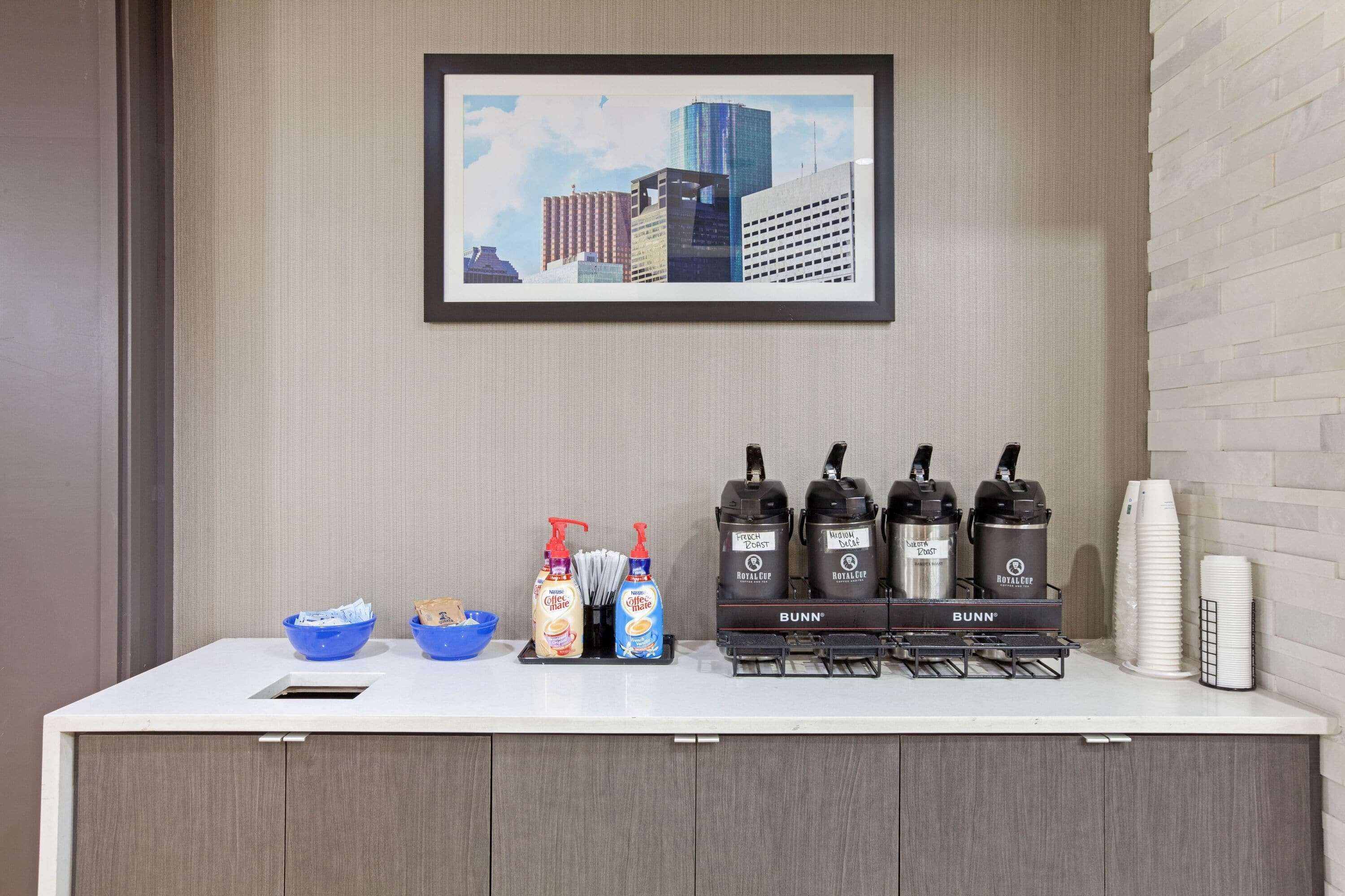 A hotel reception area with a framed cityscape picture, a coffee machine, and a counter with bowls and condiments.