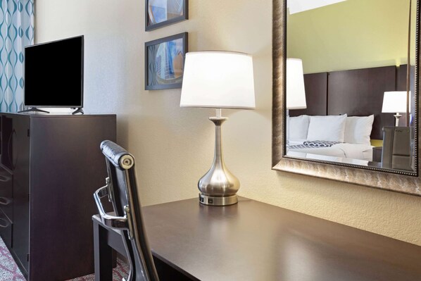 Habitación de hotel con escritorio, lámpara, televisor y un espejo grande que refleja la cama y una mesita de noche.