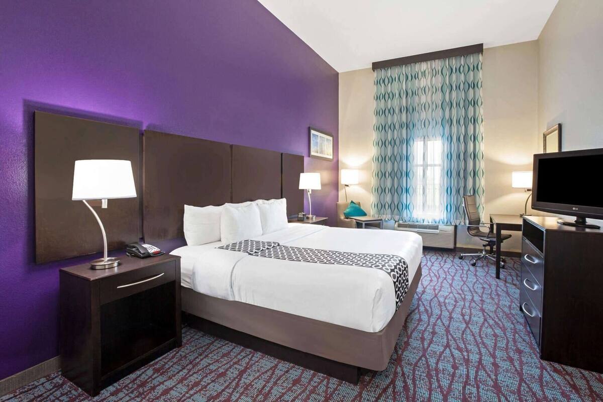 Una habitación de hotel con una cama grande, mesitas de noche con lámparas, un escritorio con televisor y una ventana con cortinas estampadas.