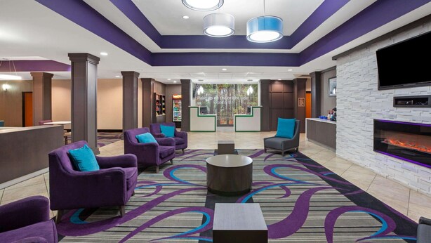 A hotel lobby with purple armchairs, a patterned carpet, a fireplace, and a TV mounted on the wall.