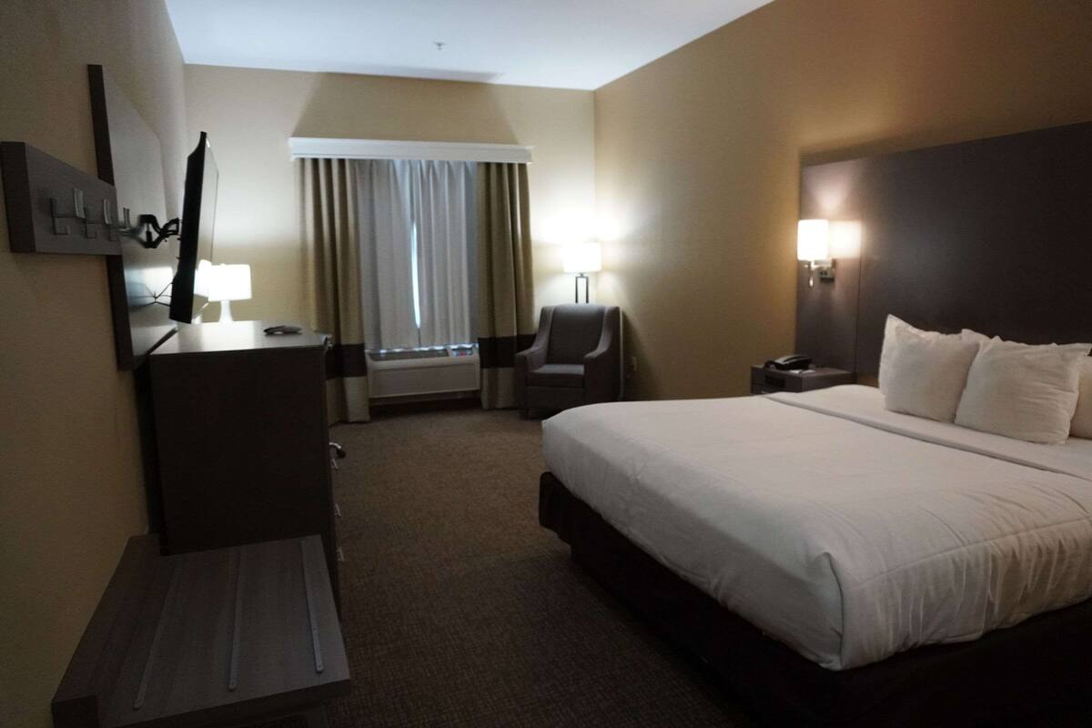 A hotel room with a large bed, a desk with a chair, a desk lamp, a chair, a mirror, and a window with curtains.