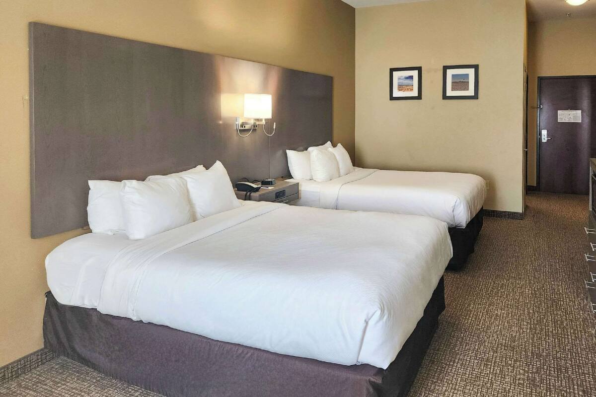 A hotel room with two beds, a headboard, a nightstand, a wall-mounted lamp, and two framed pictures on the wall.