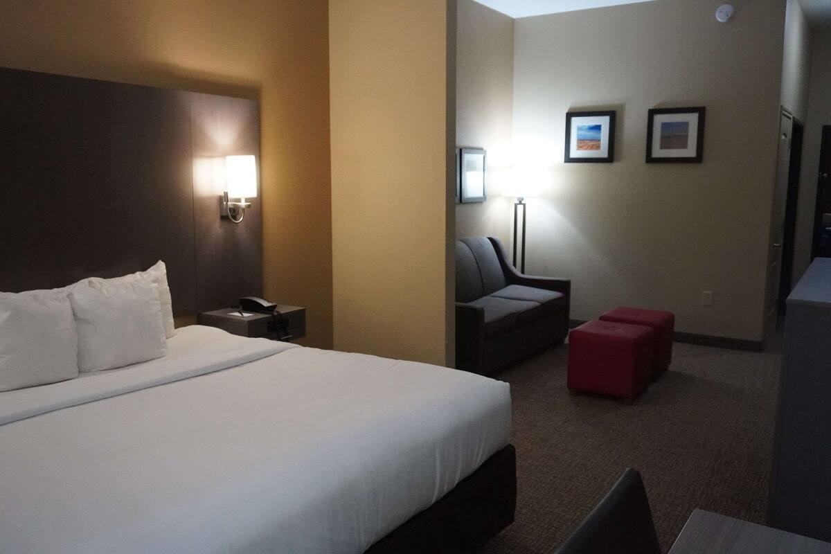 A hotel room with a large bed, a nightstand, a desk, a chair, and a red ottoman. There are framed pictures on the wall and a lamp on the desk.