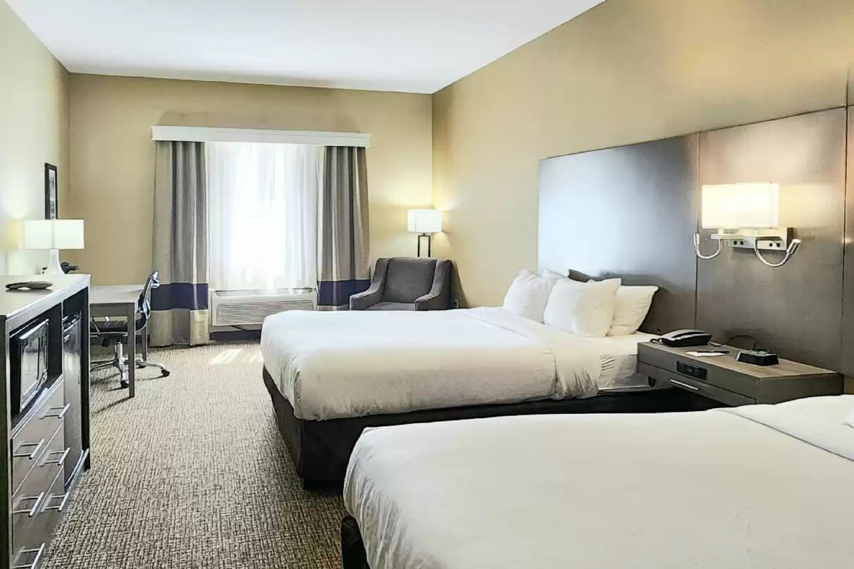 A hotel room with two beds, a desk, a chair, a lamp, and a window with curtains.