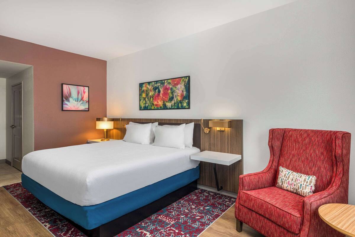 Una habitación de hotel con una cama grande, un sillón rojo, una mesita y dos lámparas de pared.