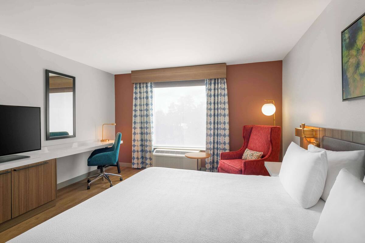 Habitación de hotel con una cama grande, un escritorio con televisor de pantalla plana, un sillón rojo, una silla de oficina azul y una ventana con cortinas estampadas.