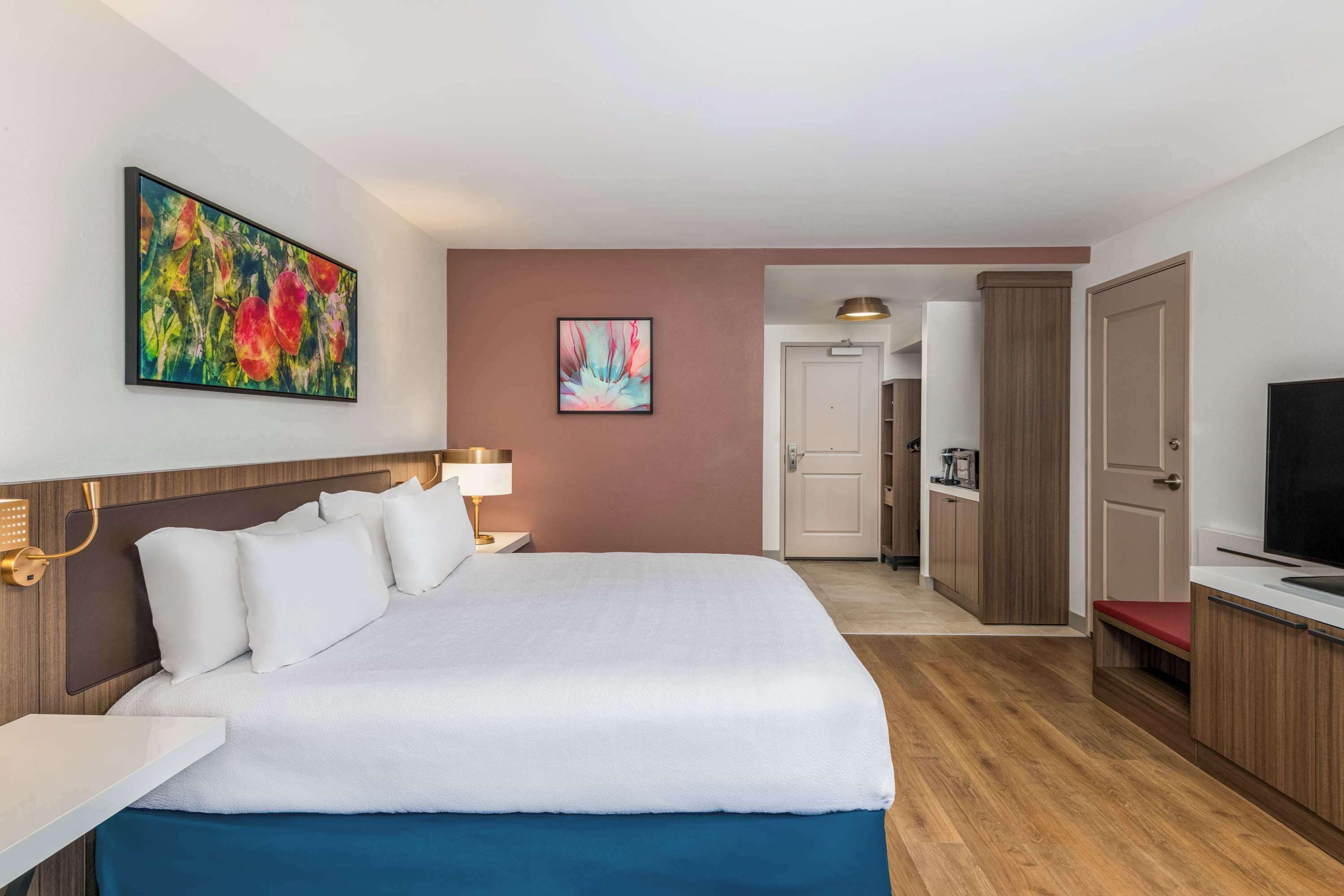 Een hotelkamer met een groot bed, een bureau, een televisie, een kitchenette en kunstwerken aan de muur.