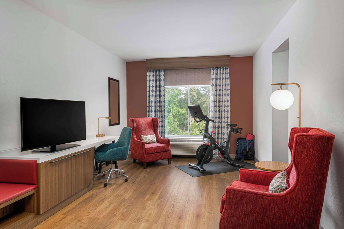 Un salón moderno con televisor de pantalla plana, sillones rojos, una silla de oficina color turquesa, una bicicleta estática y suelo de madera.