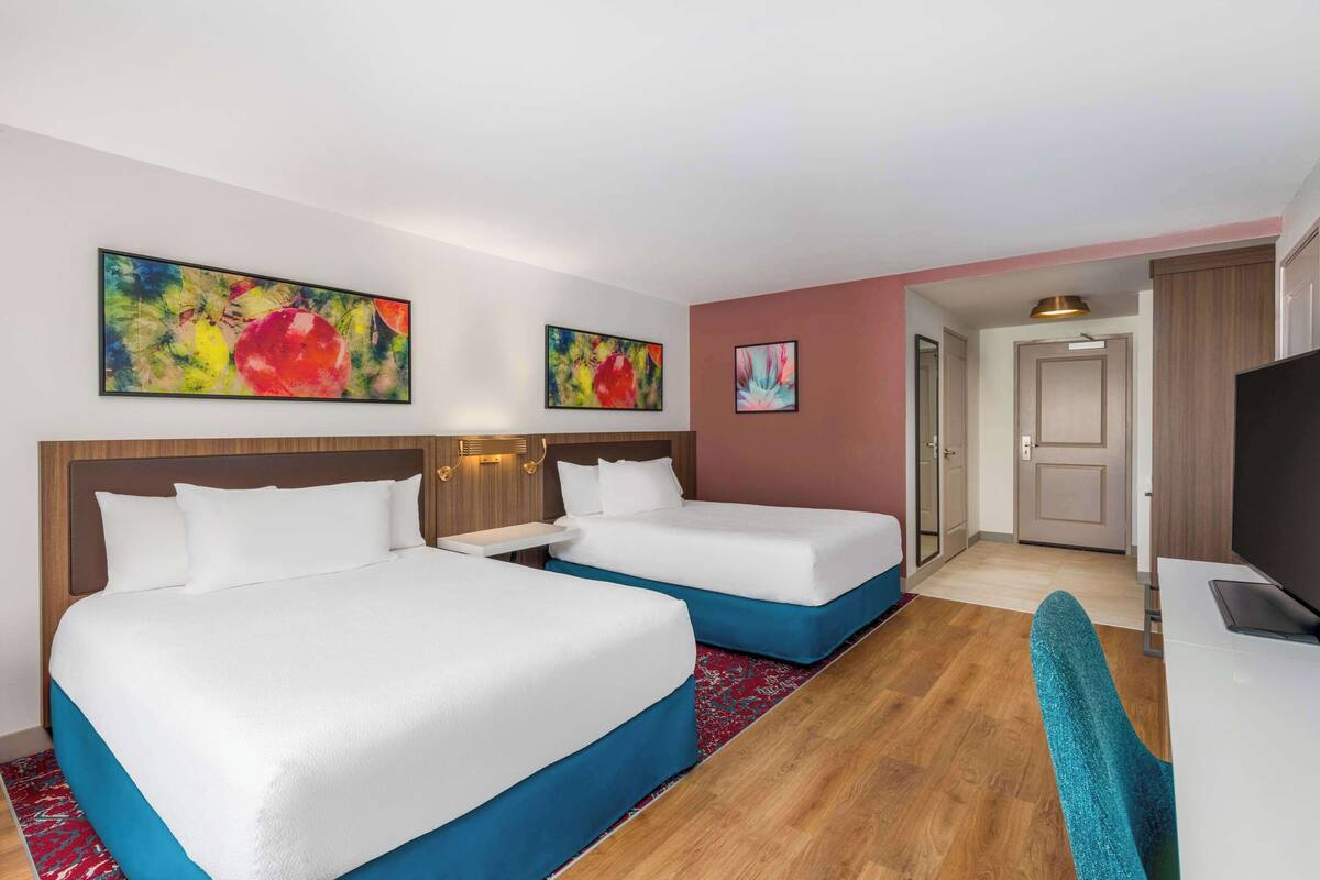 Habitación de hotel con dos camas, un escritorio, un televisor y obras de arte en las paredes.