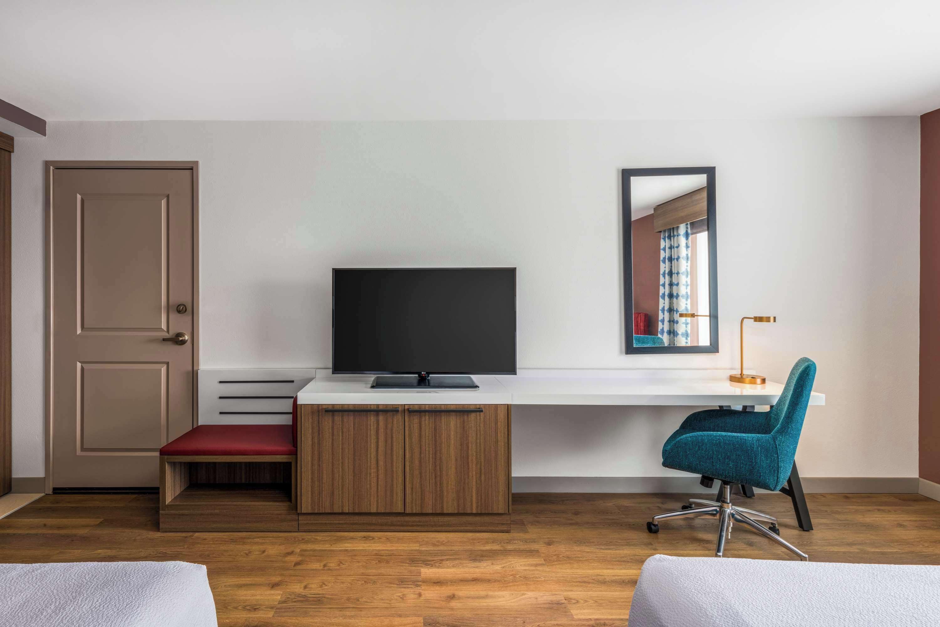 Une pièce moderne avec un plancher en bois, un bureau doté d’un téléviseur à écran plat, une chaise de bureau turquoise, un banc rouge et une porte.