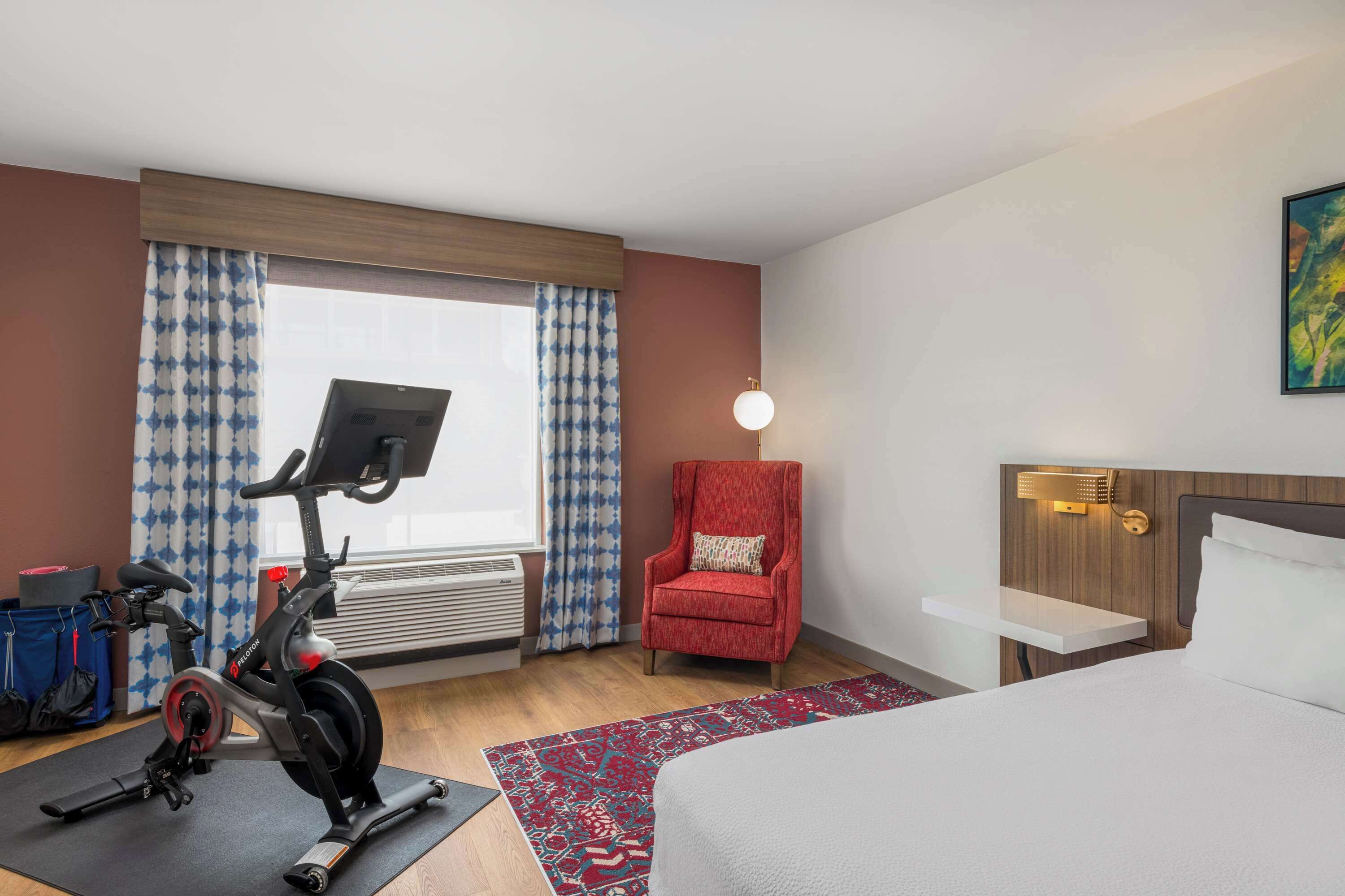 A hotel room with a bed, a red armchair, an exercise bike, a desk with a lamp, and a window with curtains.