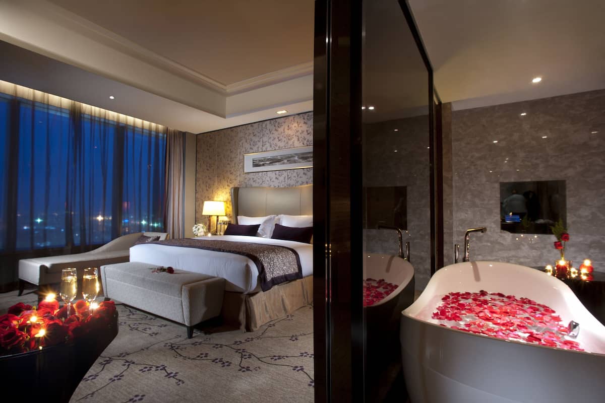 A hotel room with a large bed, a sitting area with a sofa and armchairs, a bathtub with rose petals, and a city view through the window.